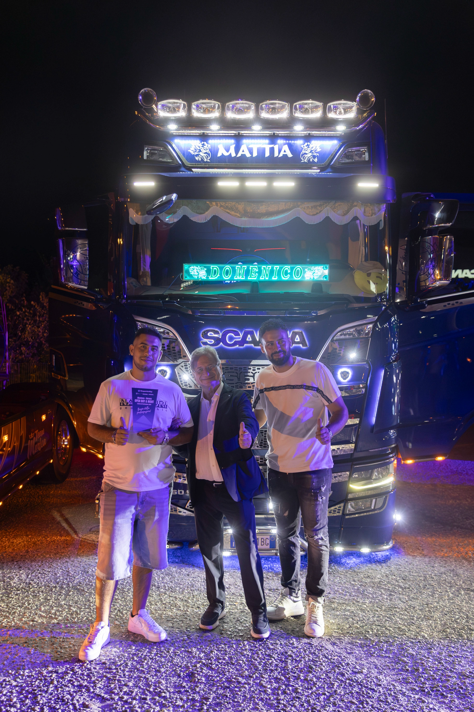 Tre uomini in posa davanti a un camion Scania decorato con luci al neon e targhe con i nomi "Mattia" e "Domenico".
