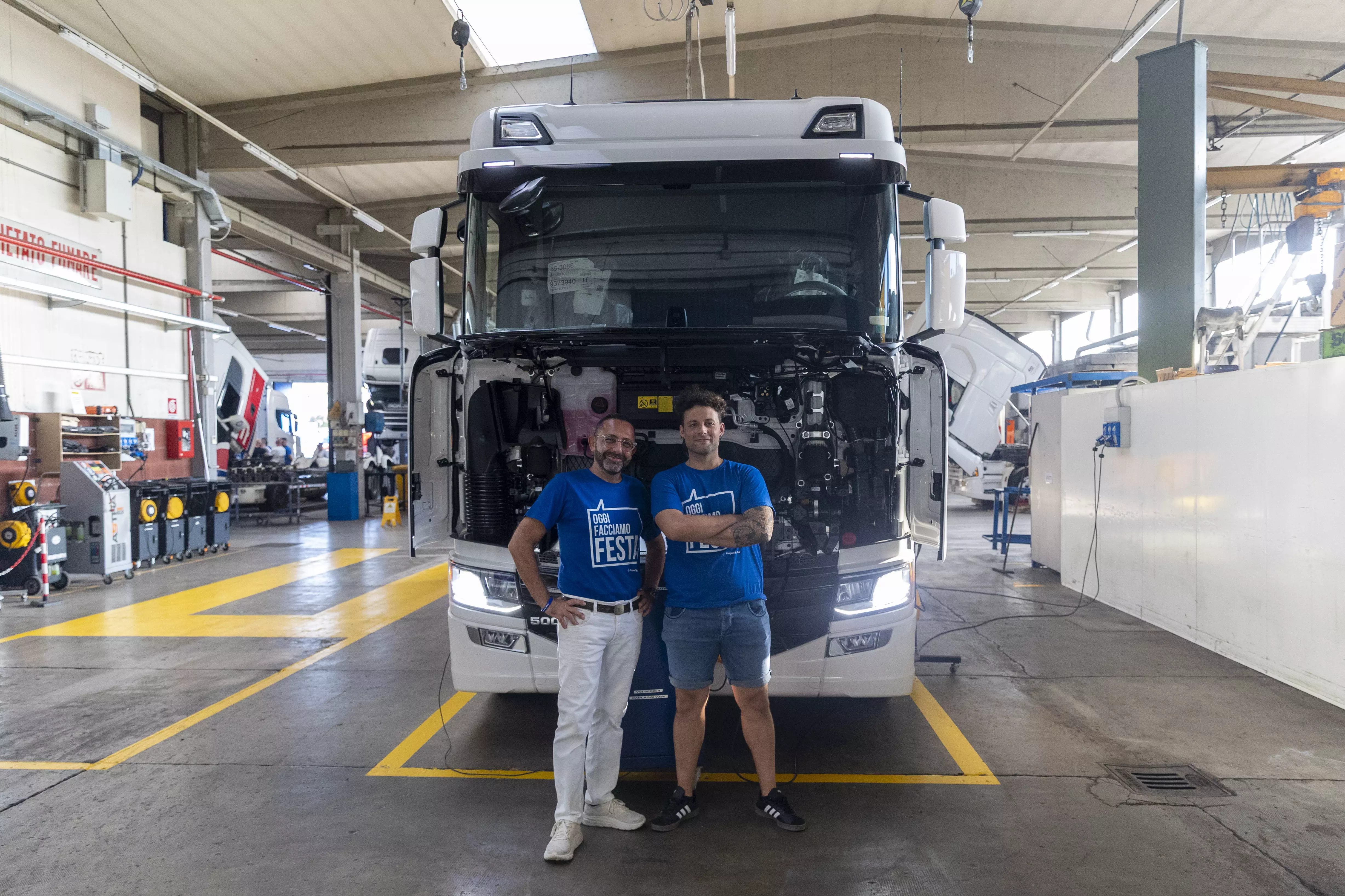 Due uomini che posano davanti a un camion in un garage con magliette blu.