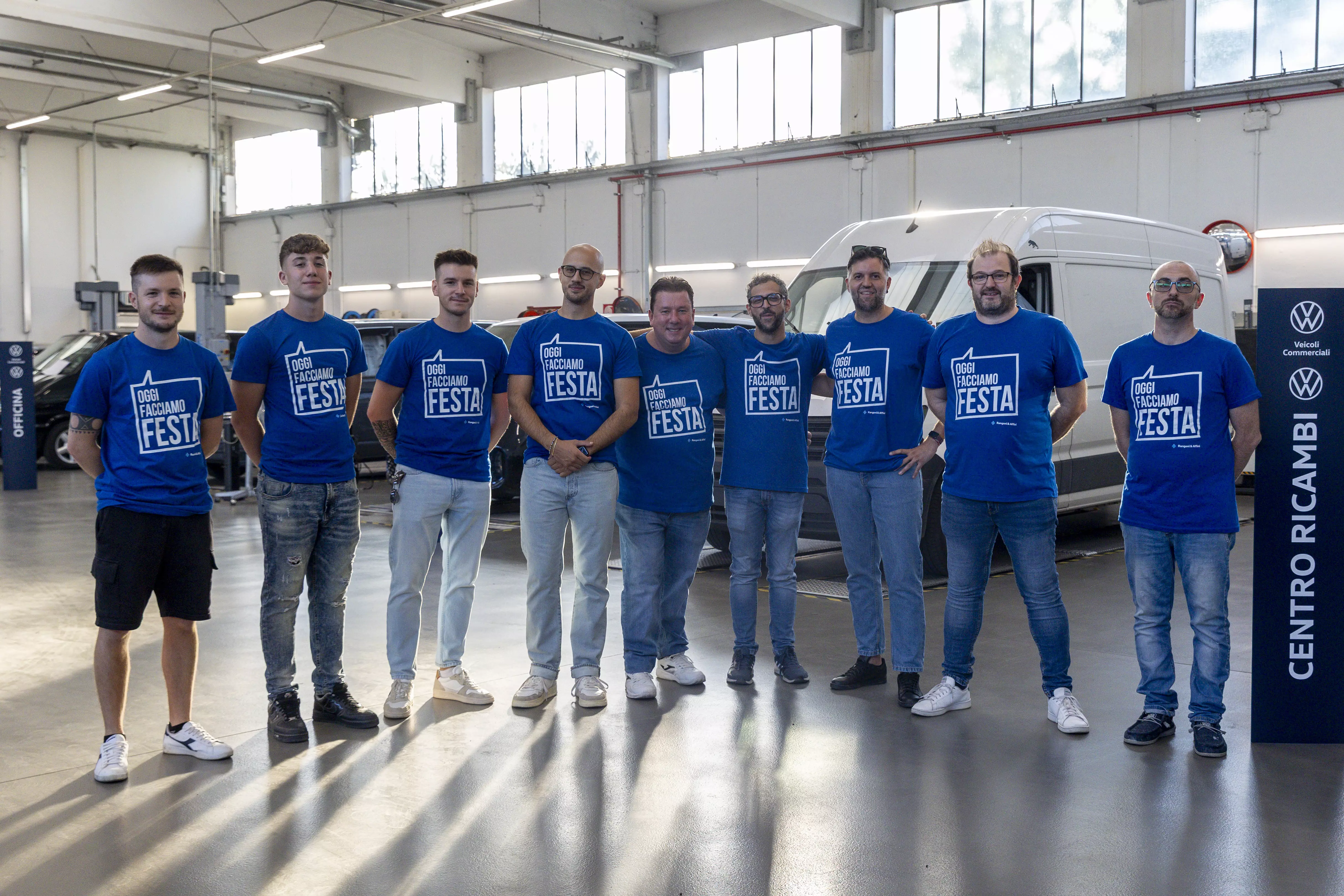 Un gruppo di nove uomini posano in un'officina meccanica con indosso magliette blu che recitano "Oggi Facciamo Festa" e vicino a un cartello Volkswagen.