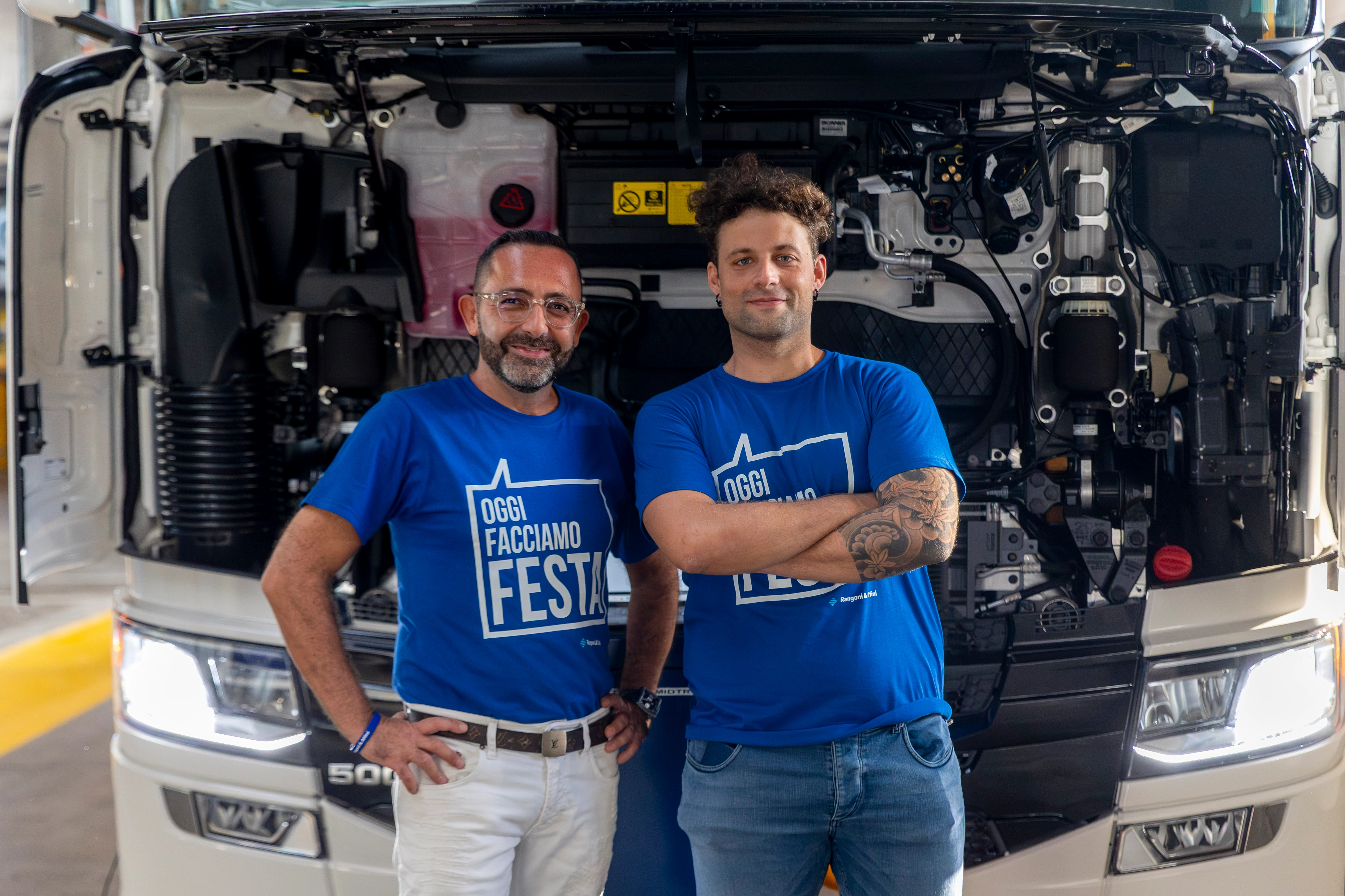 Due uomini posano davanti al motore aperto di un camion con indosso una maglietta blu con su scritto "Oggi facciamo festa".