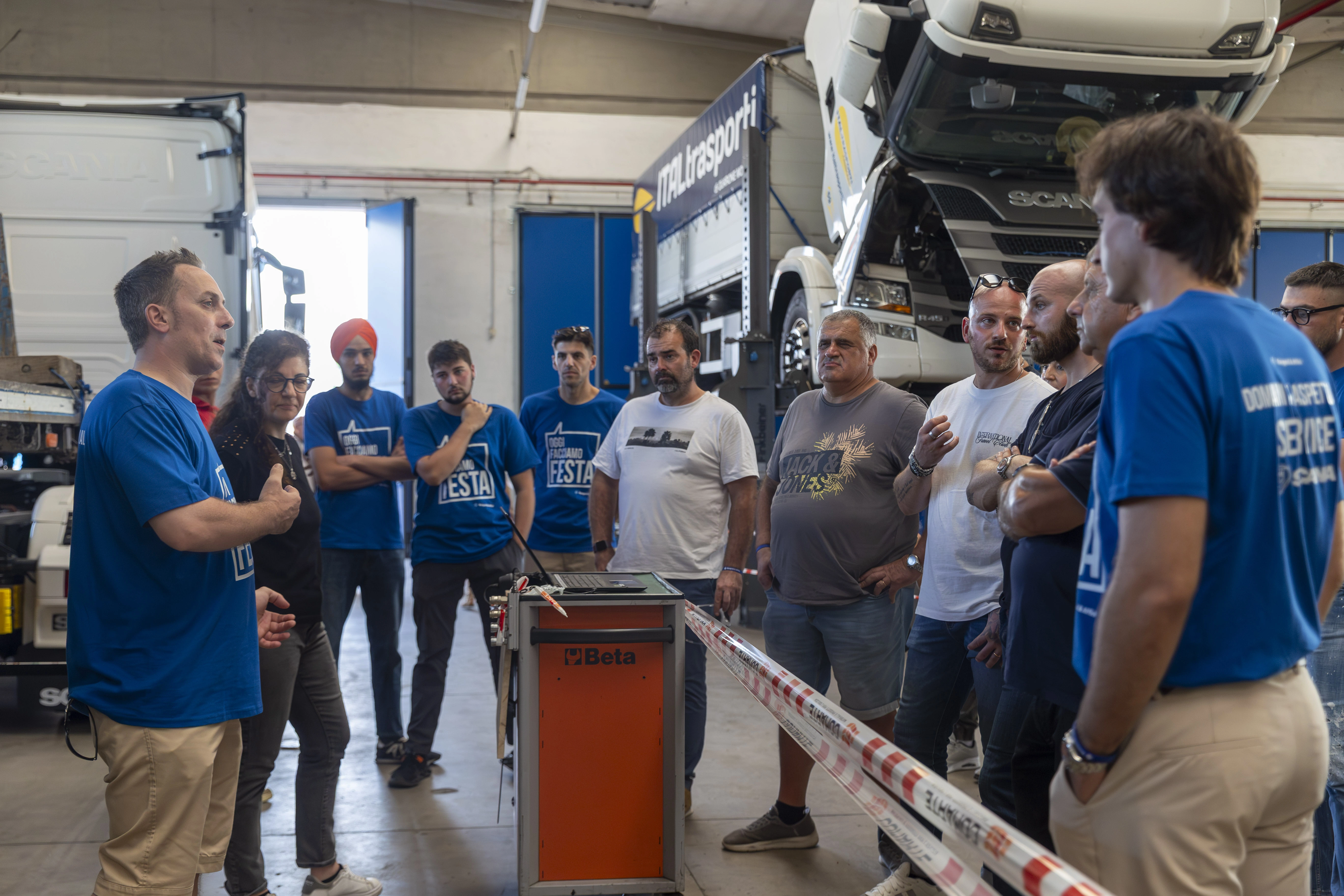 Un gruppo di persone in un'officina di riparazione di camion. Alcuni indossano magliette blu con la scritta "Oggi facciamo festa".