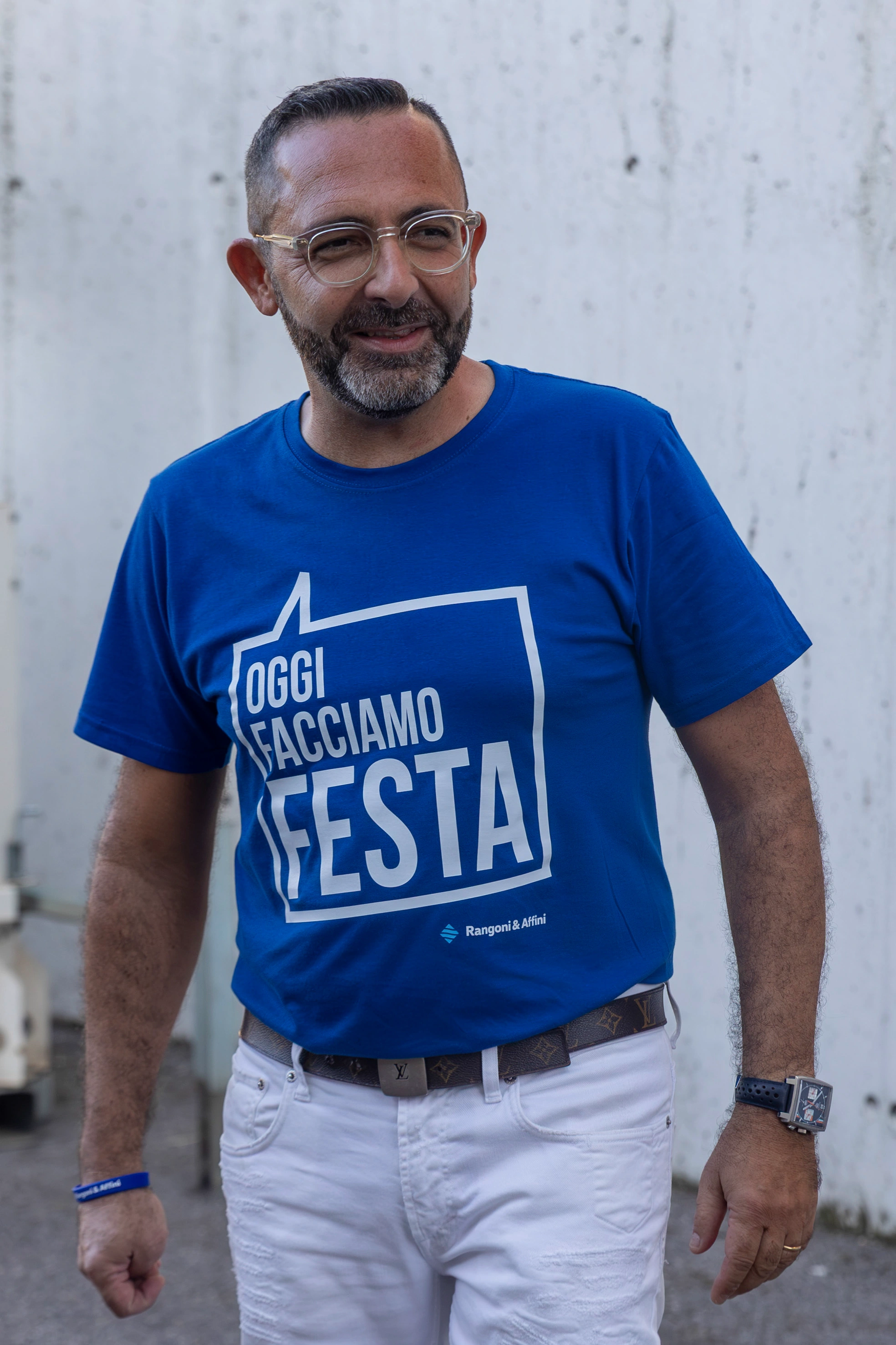Un uomo con occhiali e barba indossa una maglietta blu con la scritta "Oggi Facciamo Festa".