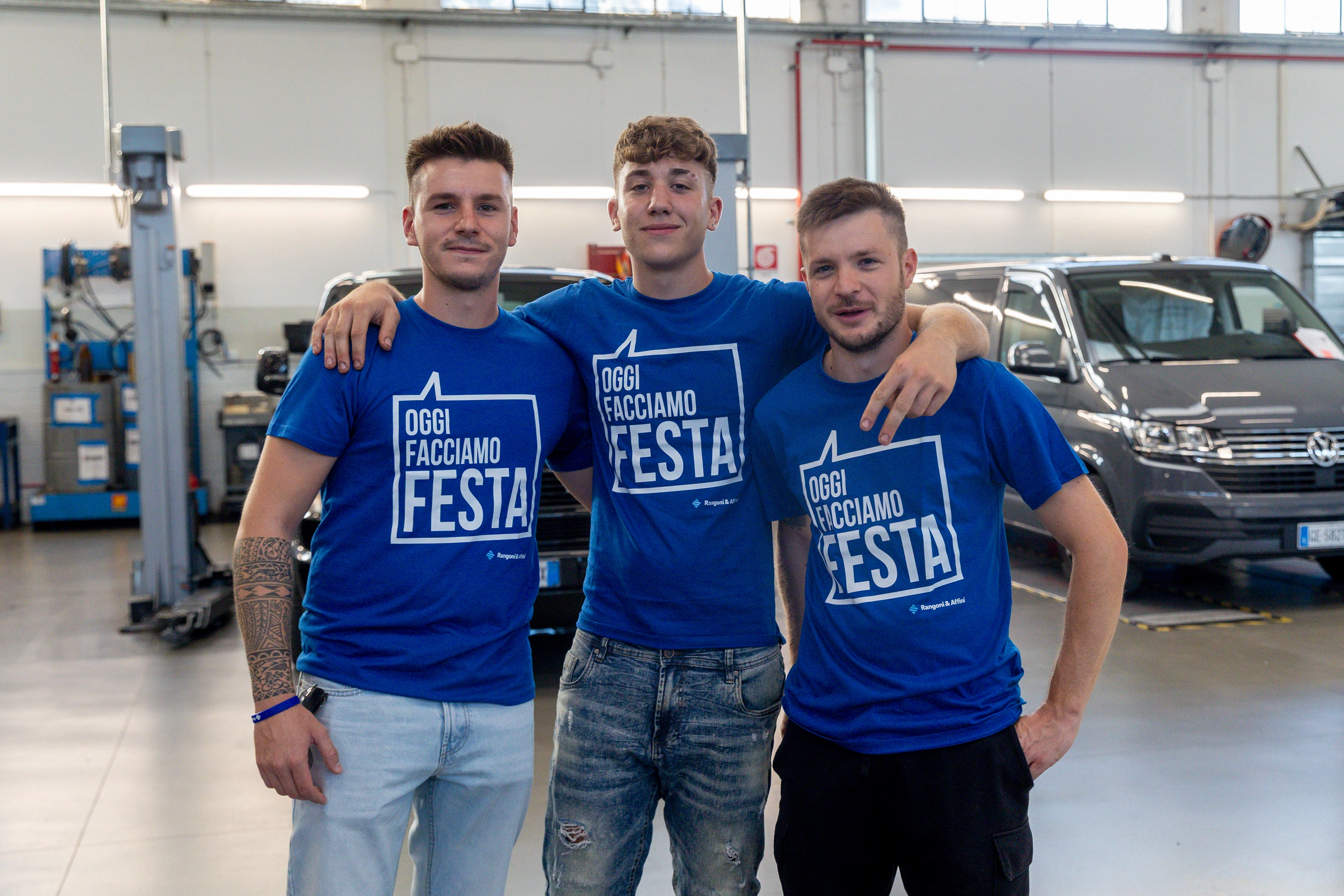 Tre giovani uomini posano sorridendo in un garage, indossando magliette blu con la scritta "Oggi Facciamo Festa".