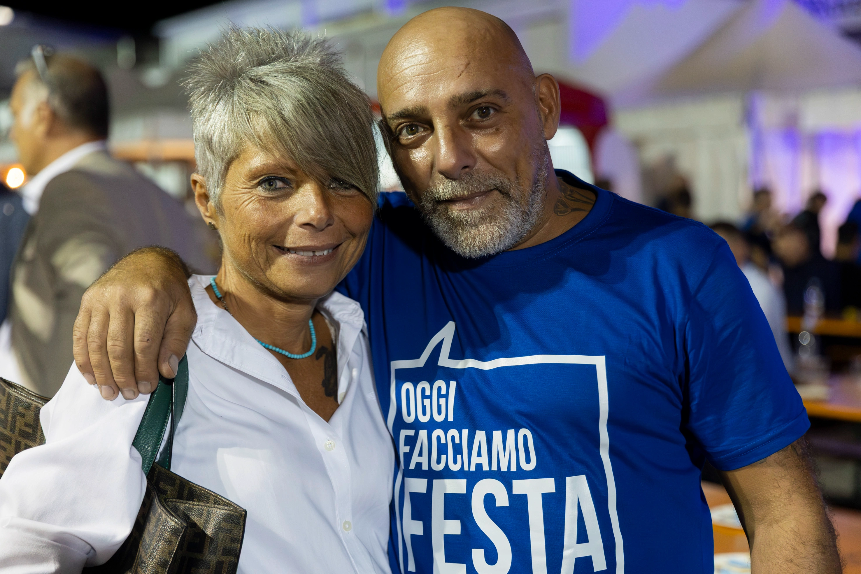 Due persone sorridono per la telecamera. L'uomo indossa una maglietta blu con la scritta "Oggi Facciamo Festa".