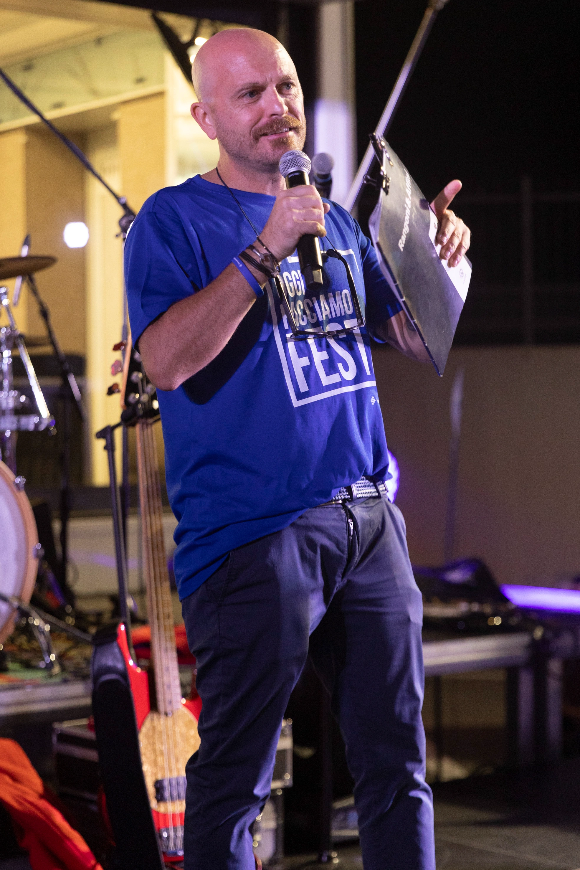 Un uomo calvo tiene un microfono e una clipboard su un palco. Sta indossando una maglietta blu.