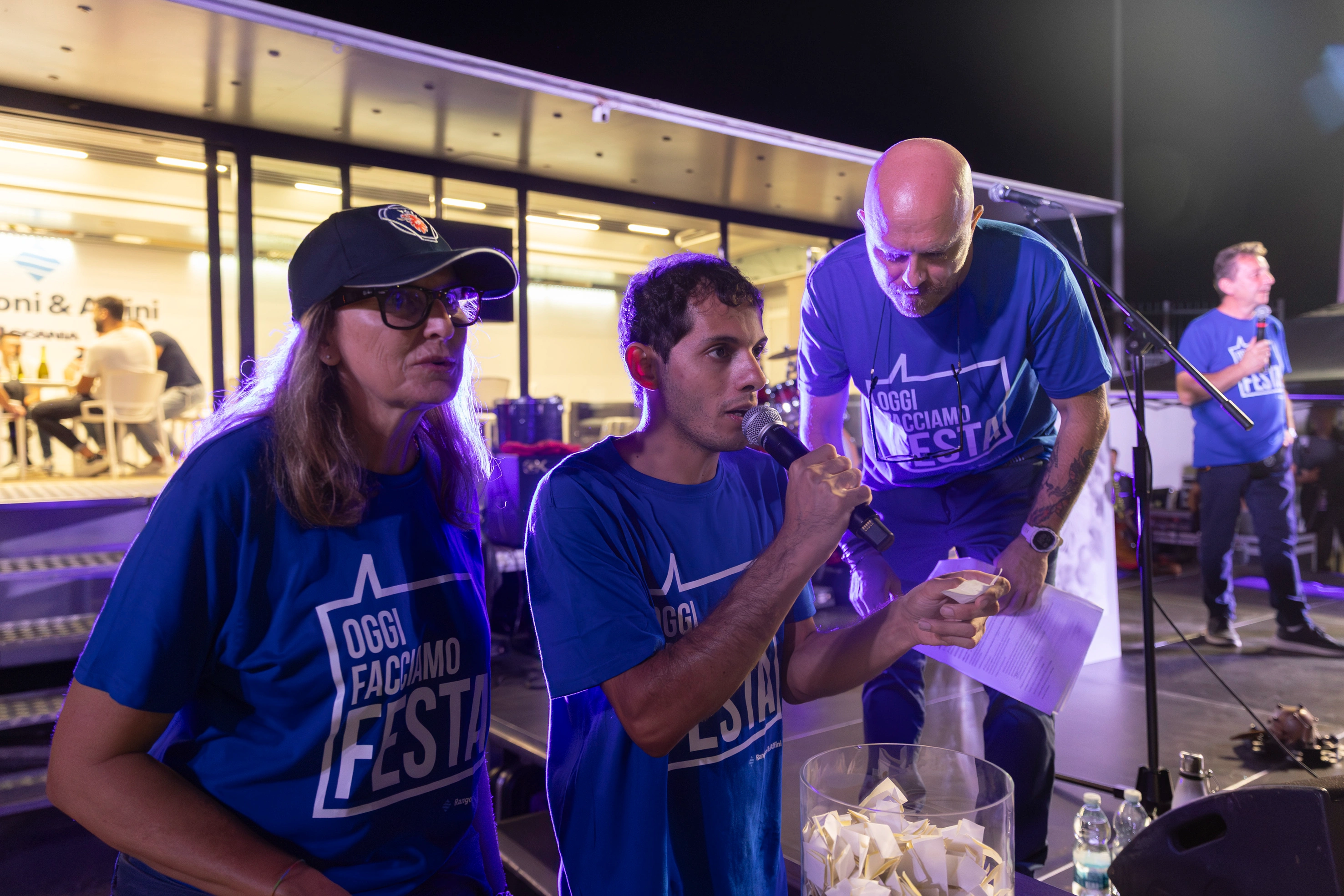 Quattro persone sul palco partecipano a un'estrazione a premi, indossando magliette blu con la scritta "Oggi Facciamo Festa".