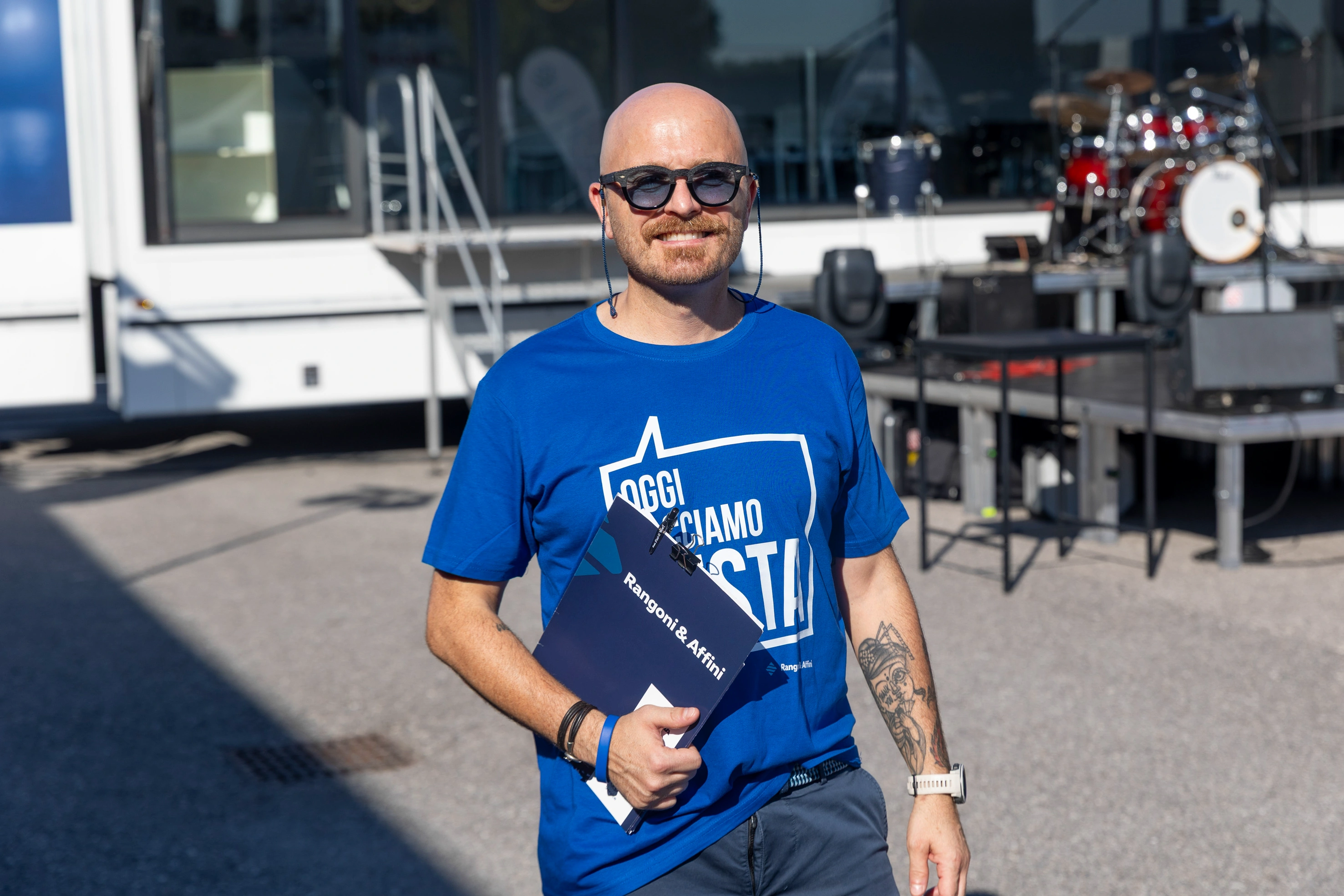 Uomo che indossa una t-shirt blu, occhiali da sole e tiene un portafoglio con la scritta "Rangoni & Affini". Sullo sfondo un palco con una batteria.