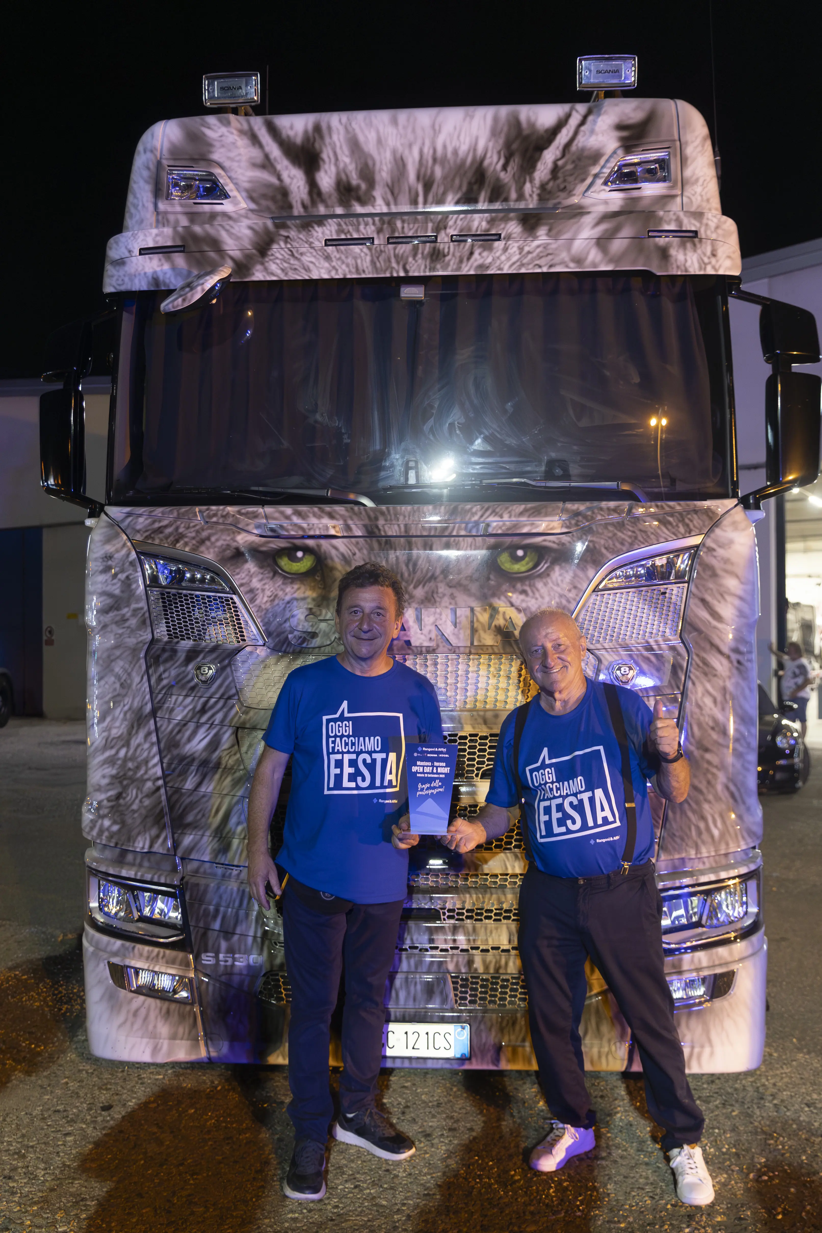 Due uomini posano davanti a un camion Scania personalizzato, indossando magliette "Oggi facciamo festa" e mostrando un premio.