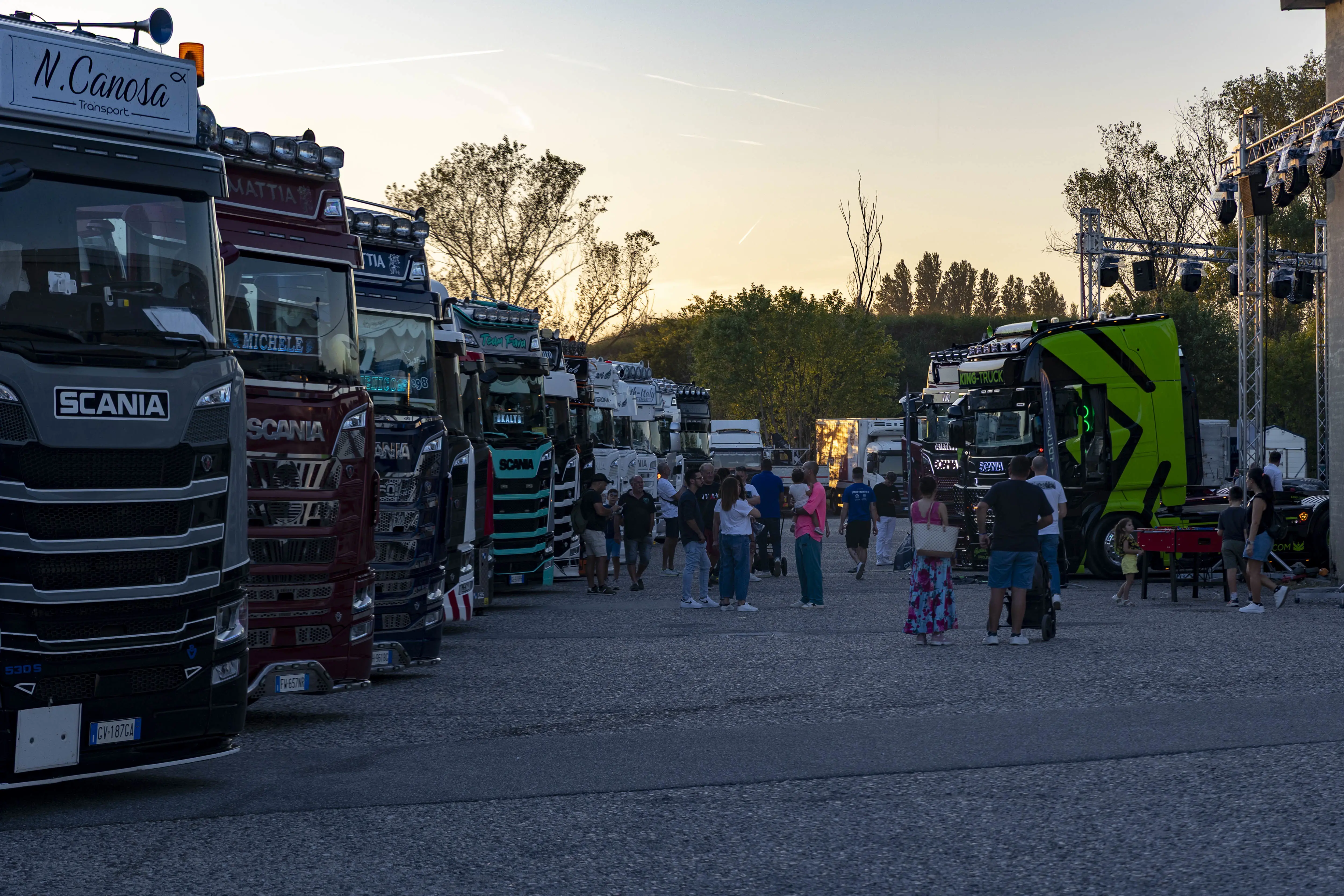 Un parcheggio affollato di camion Scania con persone che si godono un evento al tramonto.