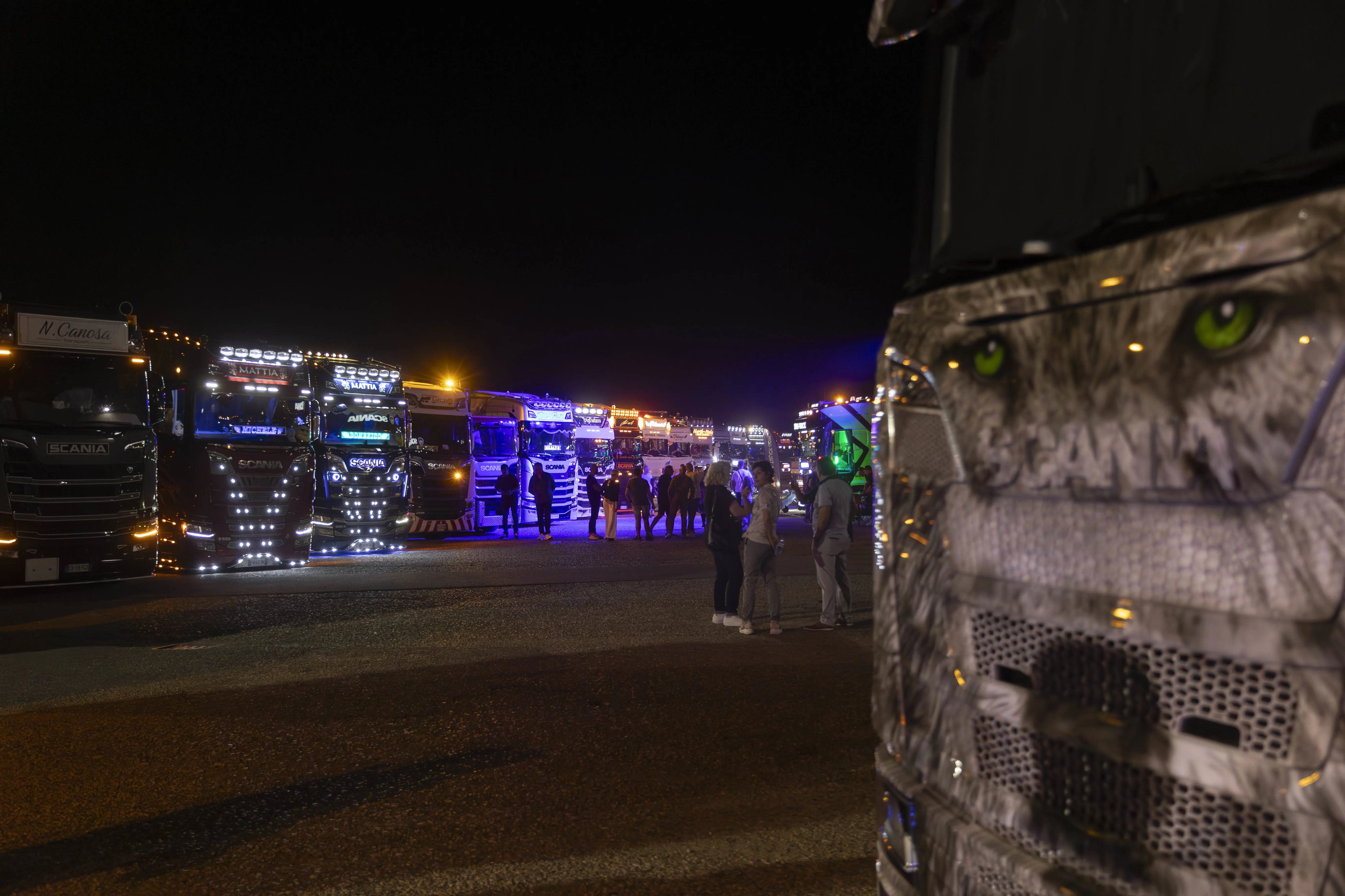 Una fila di camion Scania pesantemente modificati attira l'attenzione della folla in questa foto notturna. Il camion in primo piano è dipinto con una faccia di lupo.