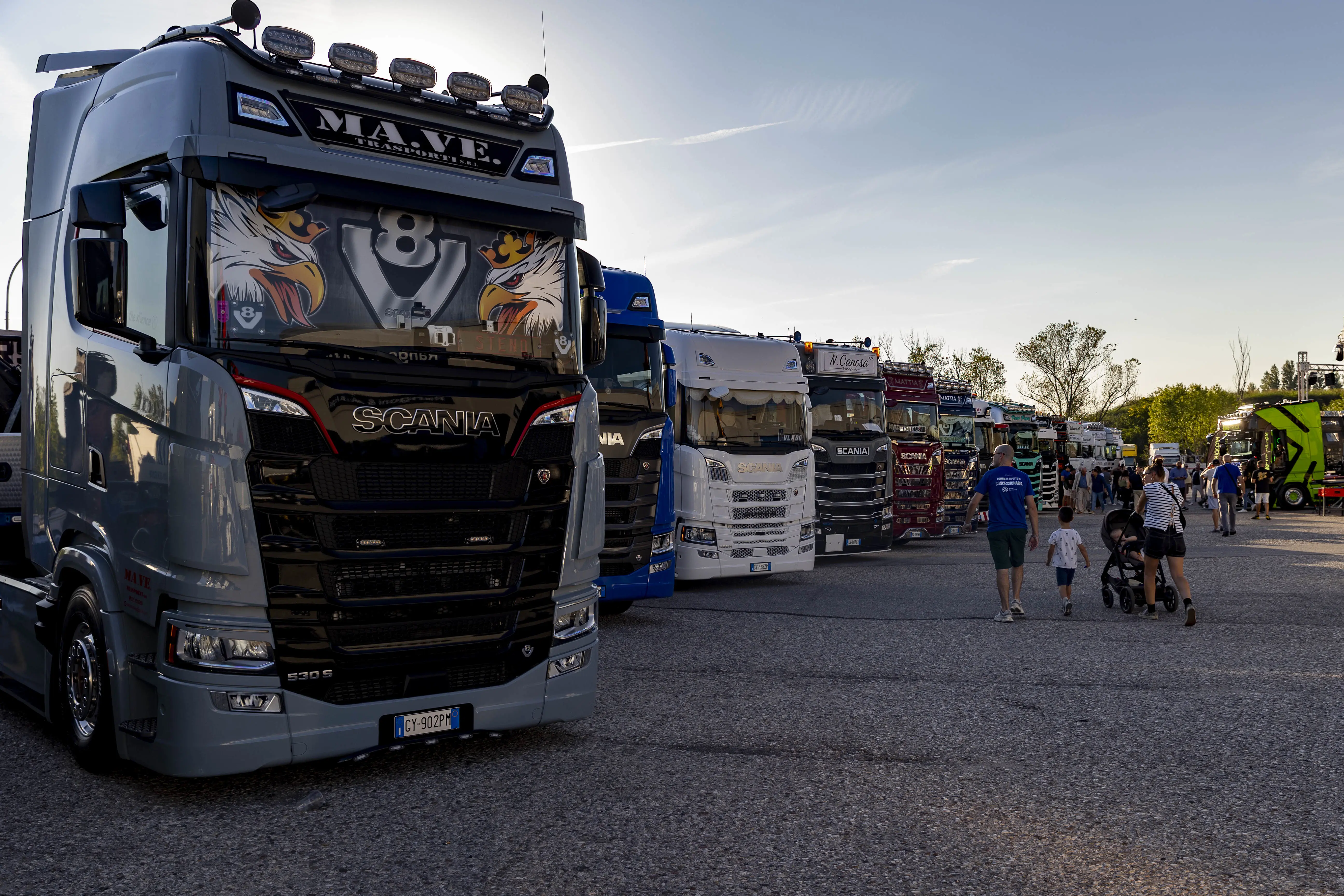 Camion Scania parcheggiati in fila in un'area di pavimentazione con persone che camminano nelle vicinanze.