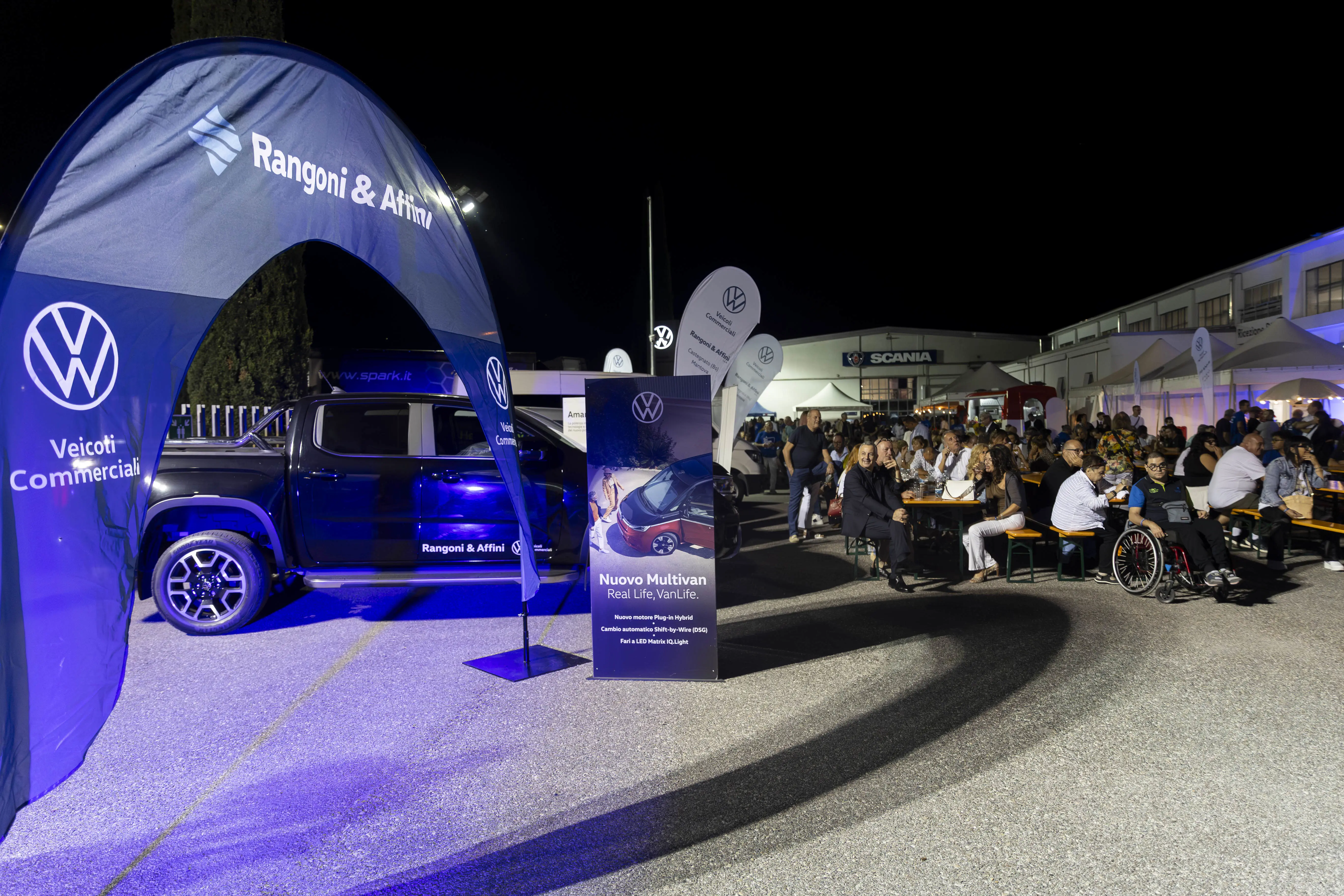 Un evento notturno con una Volkswagen Amarok in mostra, circondata da persone sedute a tavoli con cartelli e banner.