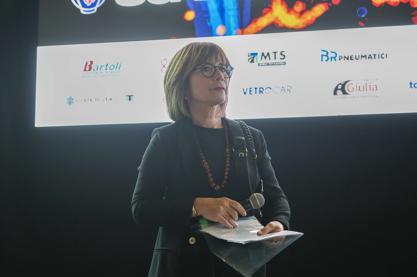 Una donna con gli occhiali tiene un microfono e dei documenti davanti a uno schermo.