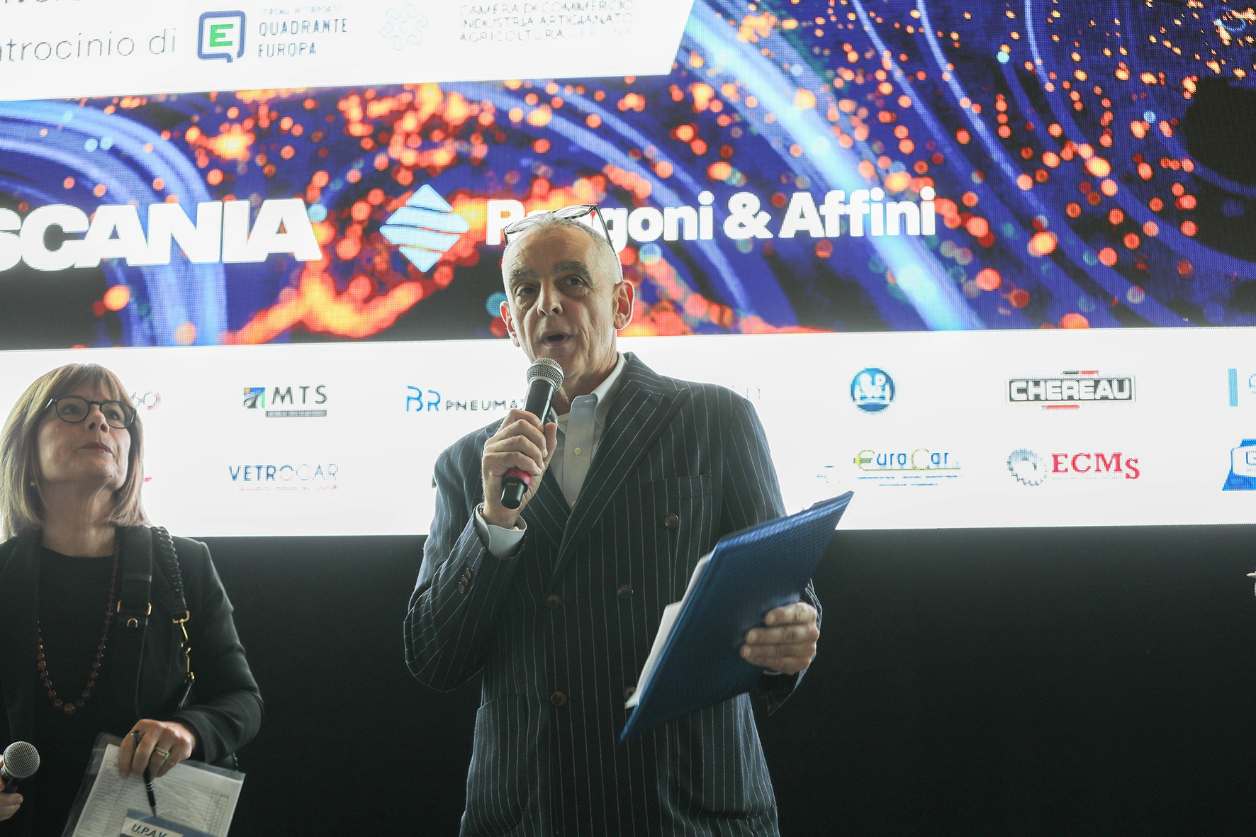 Un uomo in giacca gessata parla a un microfono davanti a uno schermo con loghi aziendali.