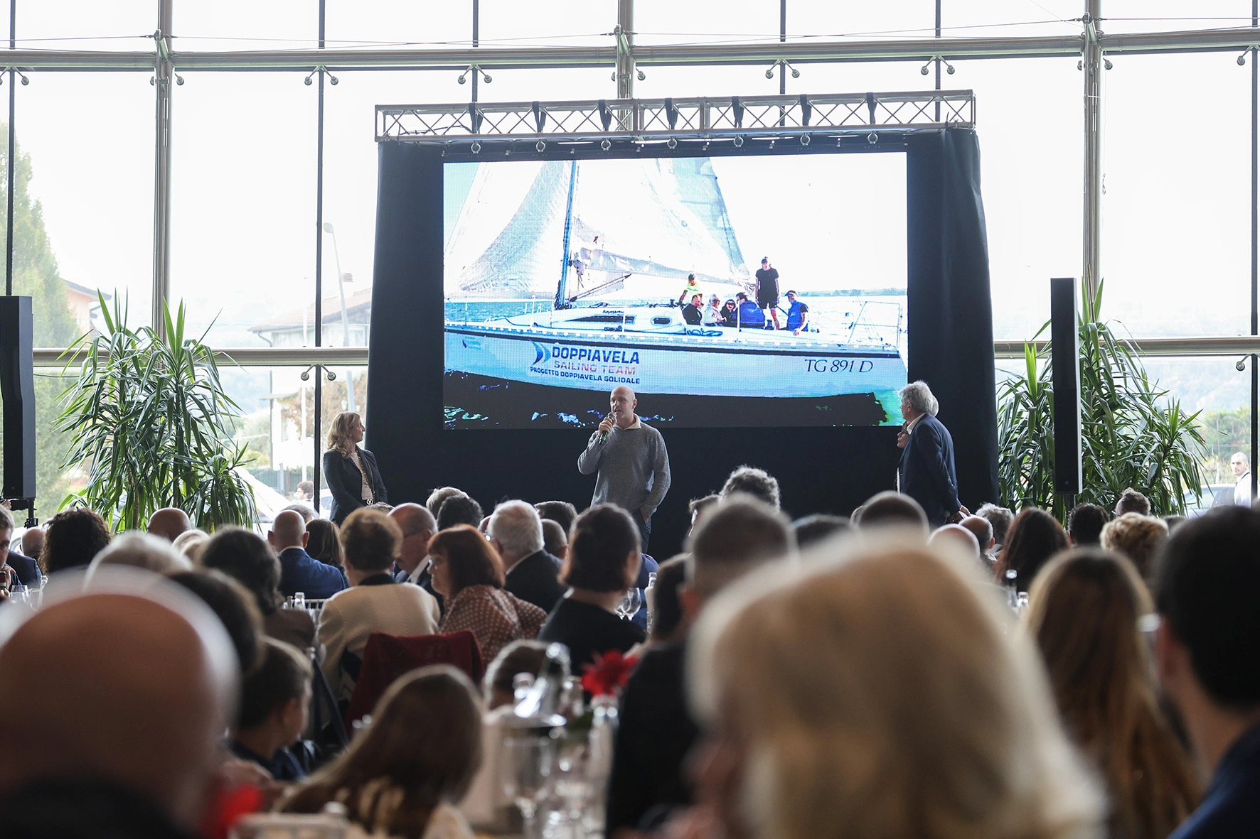 Persone riunite a una conferenza con un oratore in piedi davanti a uno schermo che mostra una barca a vela.