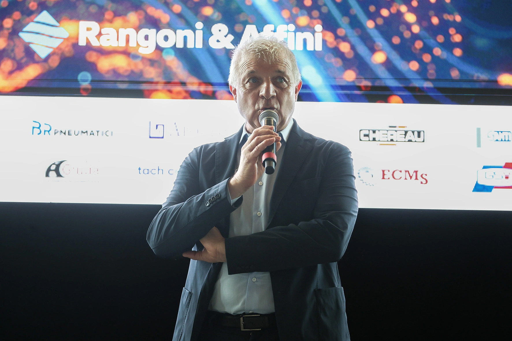 Un uomo dai capelli grigi tiene un microfono con le braccia incrociate sullo sfondo di loghi aziendali.
