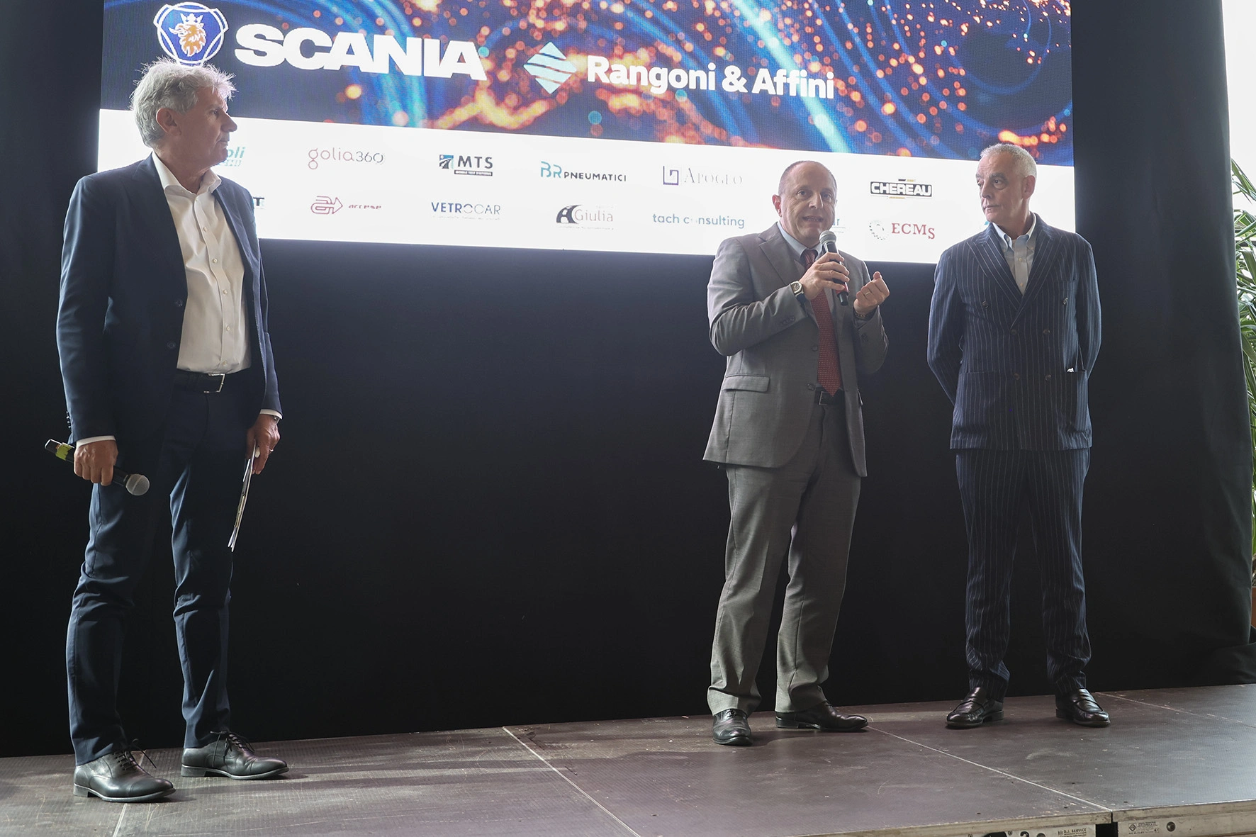 Tre uomini sul palco davanti a uno schermo con marchi.