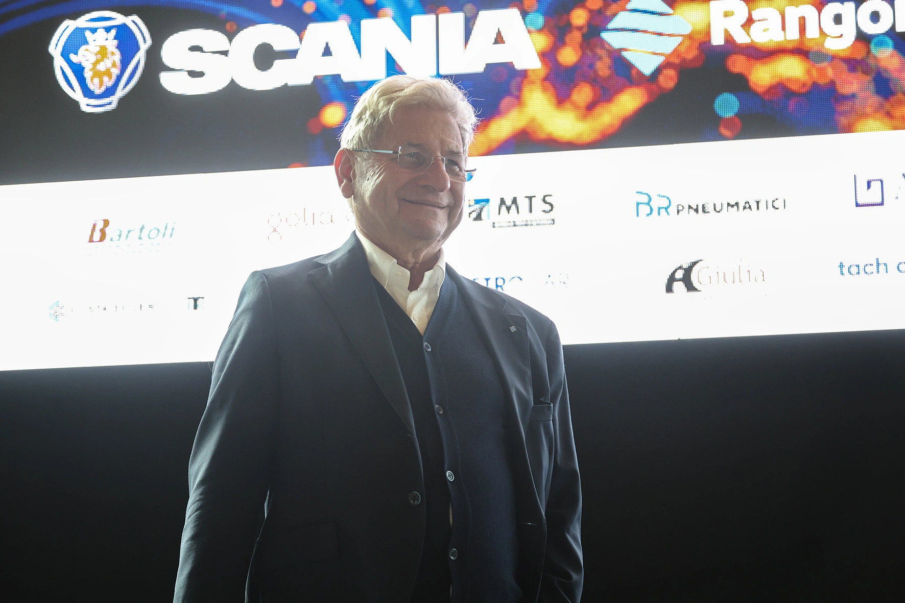 Uomo in abito blu su sfondo di loghi aziendali.