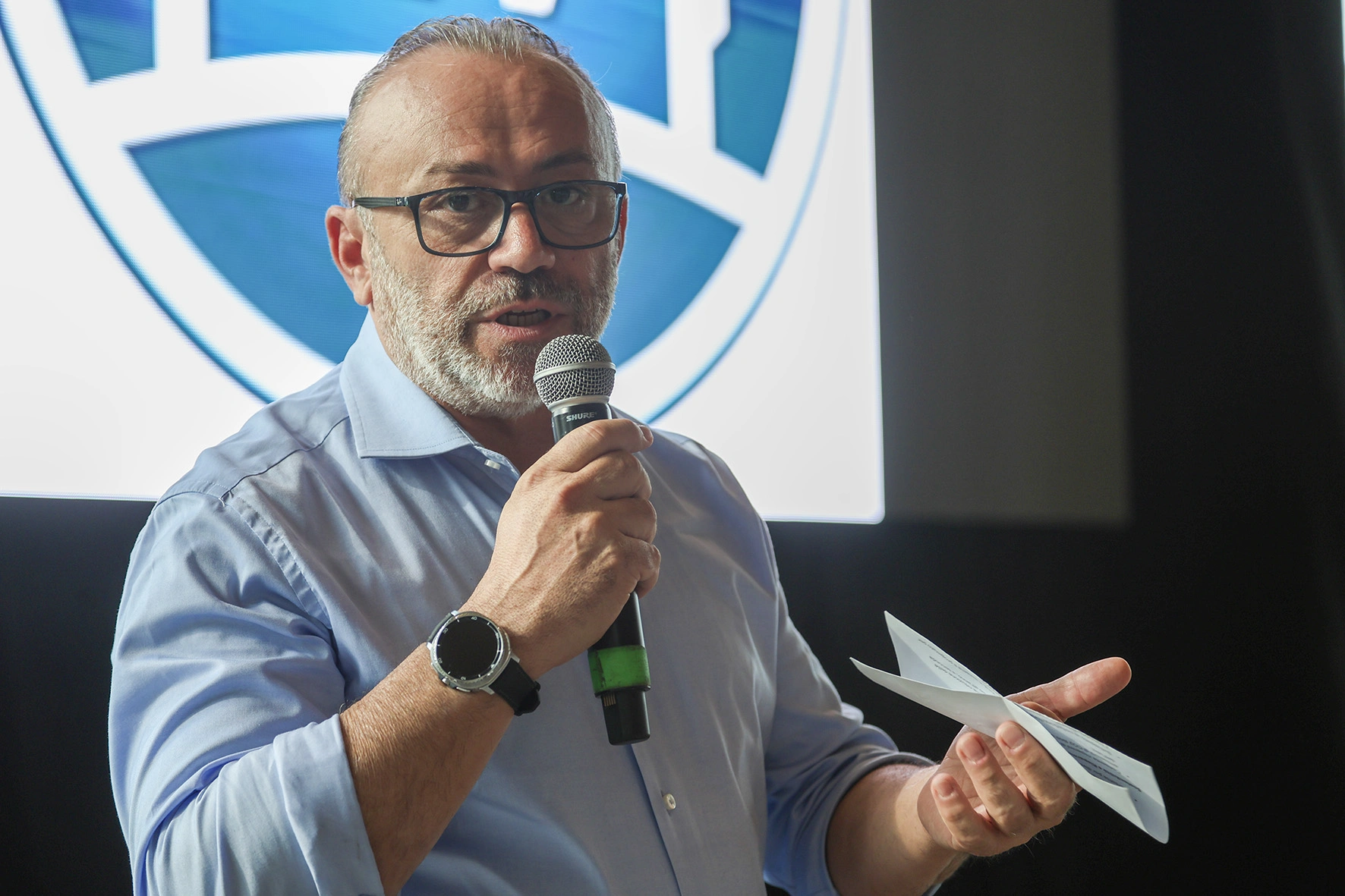 Un uomo con gli occhiali parla in un microfono e tiene in mano un foglio di carta.