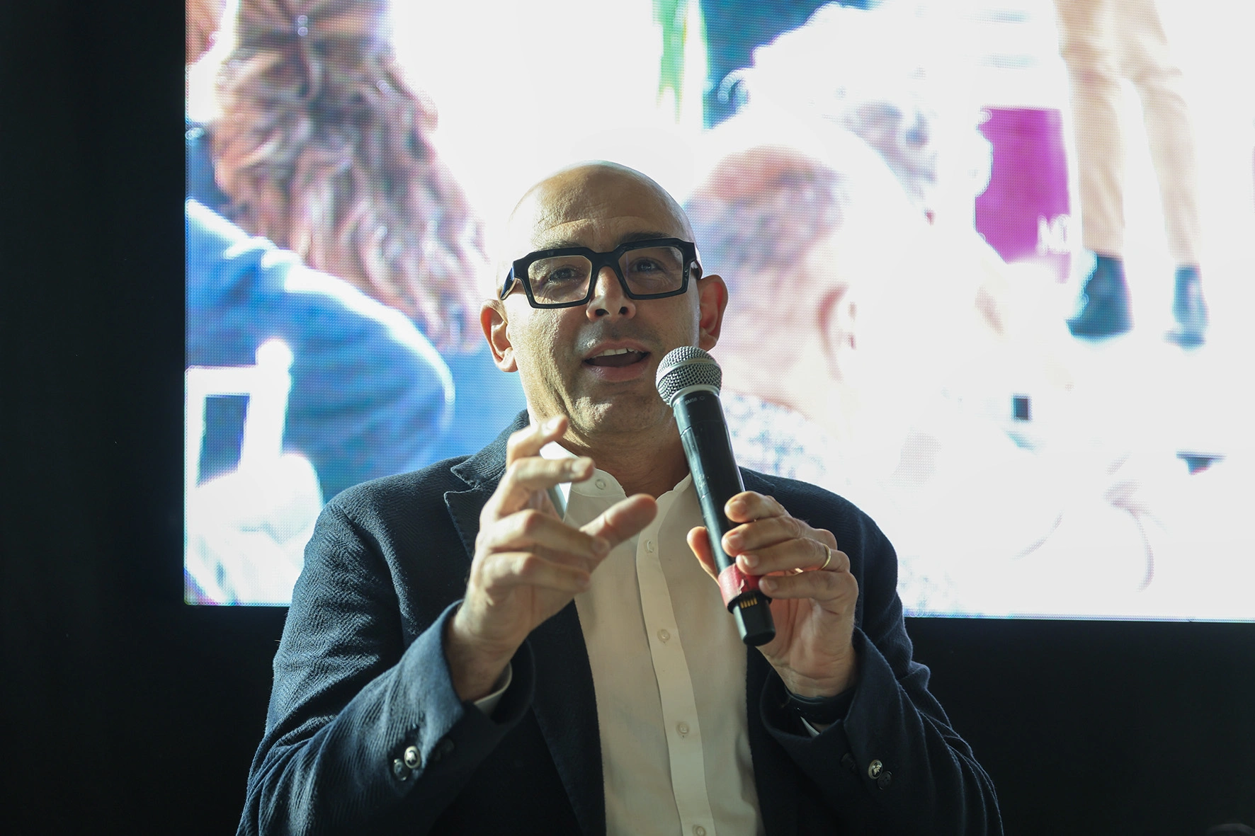 Uomo calvo che parla al microfono con gli occhiali.