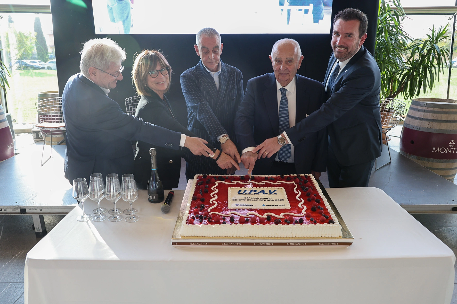 Un gruppo di persone taglia una torta per un evento UPAV. La torta è decorata con frutta e scritte.