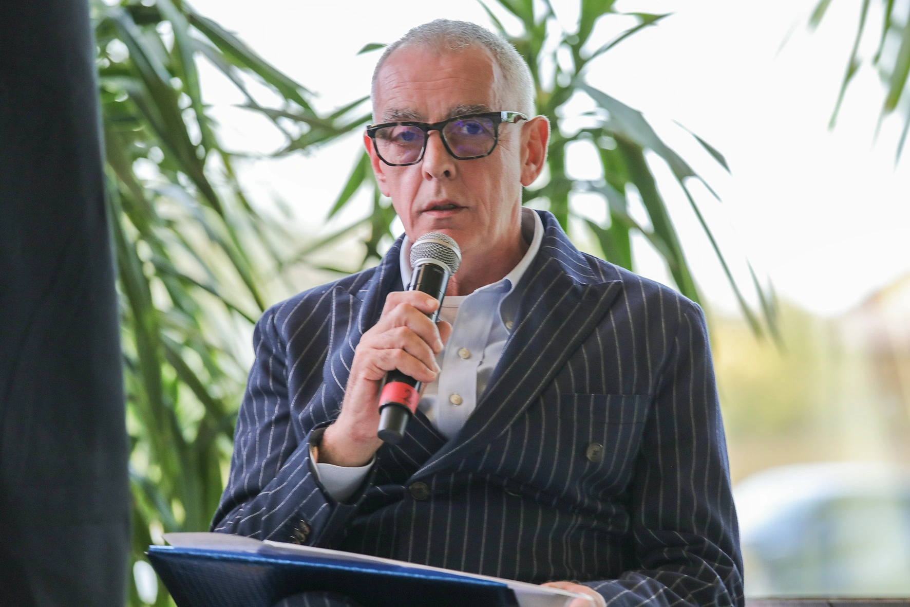 Un uomo con gli occhiali parla al microfono, indossa un abito gessato blu.