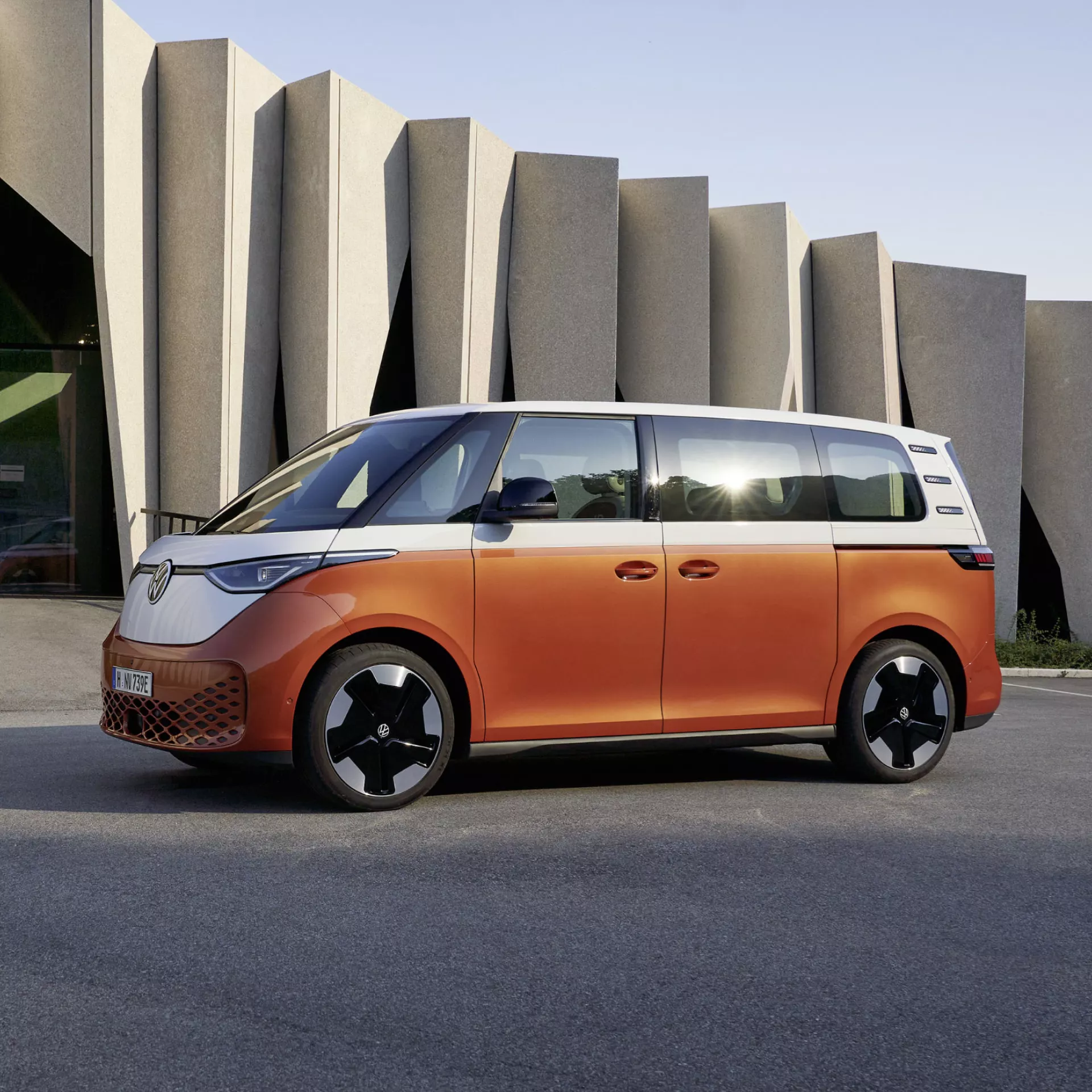 Un furgone elettrico Volkswagen ID. Buzz arancione e bianco parcheggiato fuori da un edificio moderno.