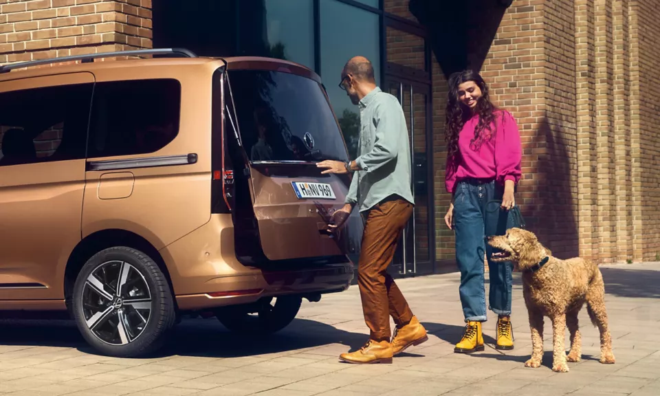 Una famiglia apre il bagagliaio del loro furgone marrone in un ambiente urbano mentre il loro cane li guarda.