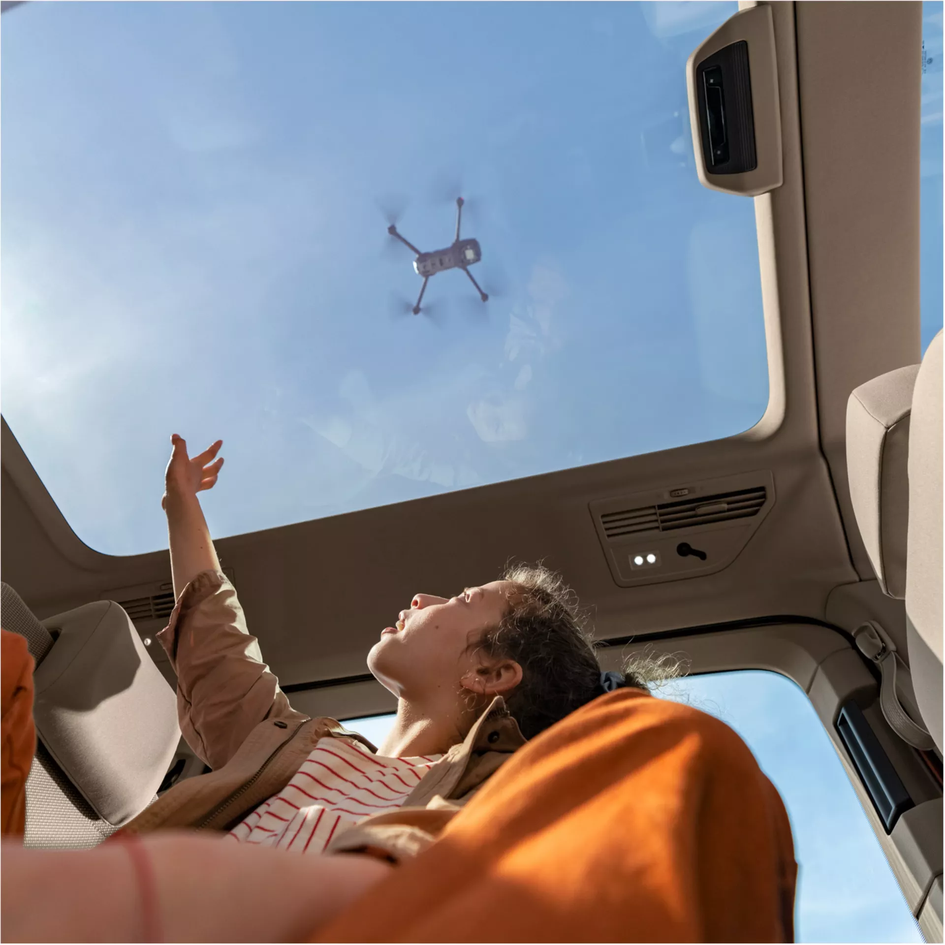 Donna distesa in auto con tetto panoramico, guarda un drone nel cielo.