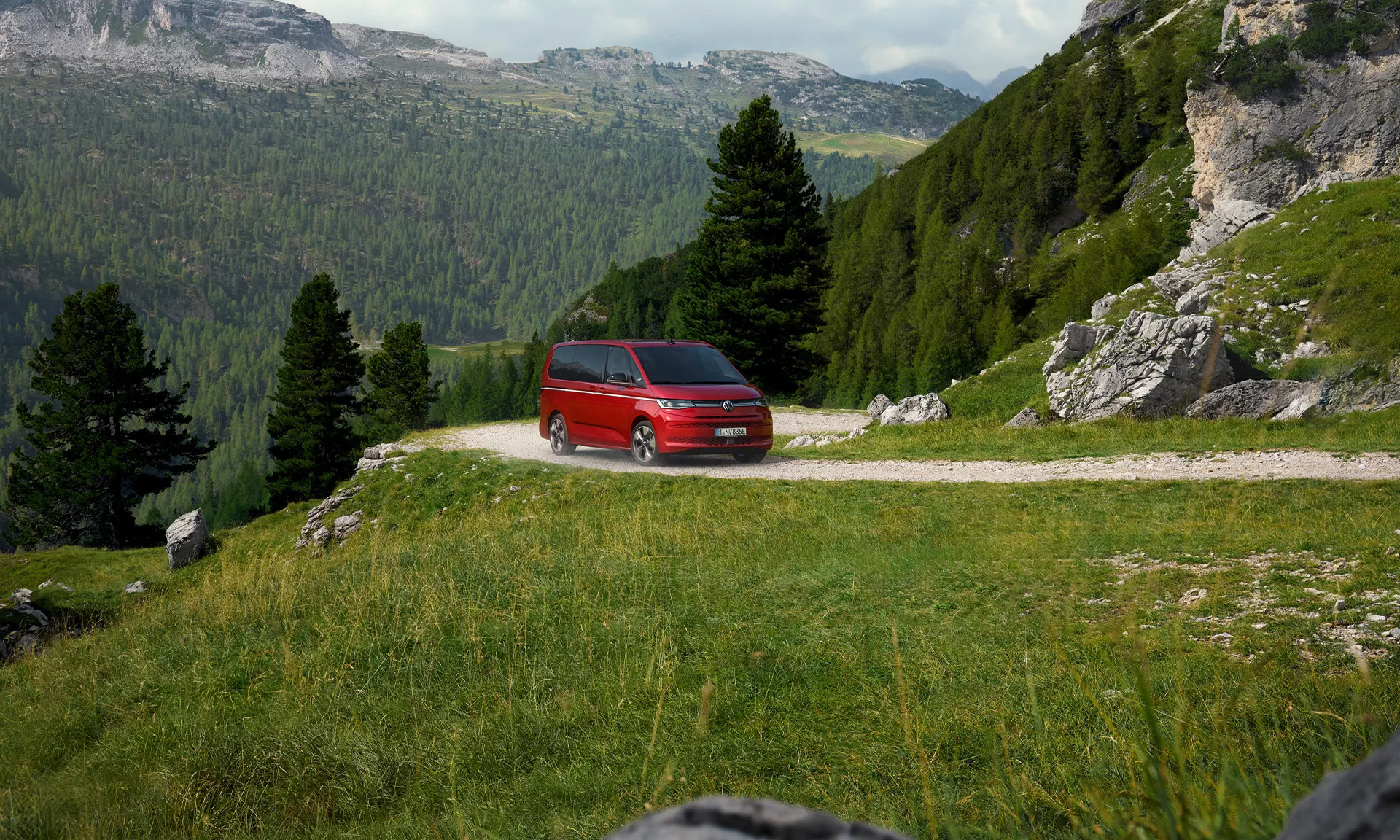 Un furgone rosso viaggia su una strada sterrata di montagna con alberi e cielo nuvoloso.