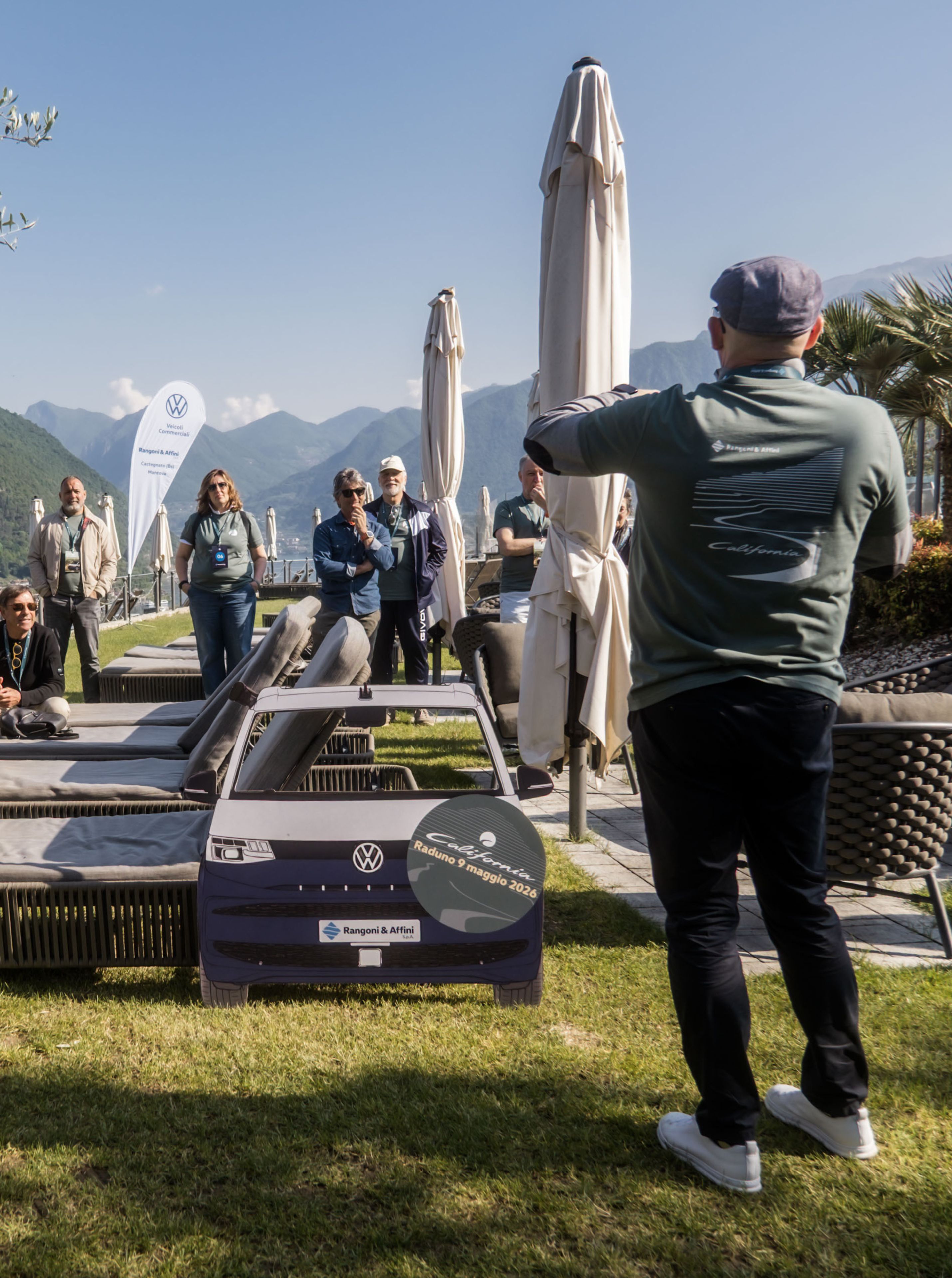 Un oratore e il pubblico a un evento Volkswagen California Veicoli Commerciali all'aperto, con montagne sullo sfondo.