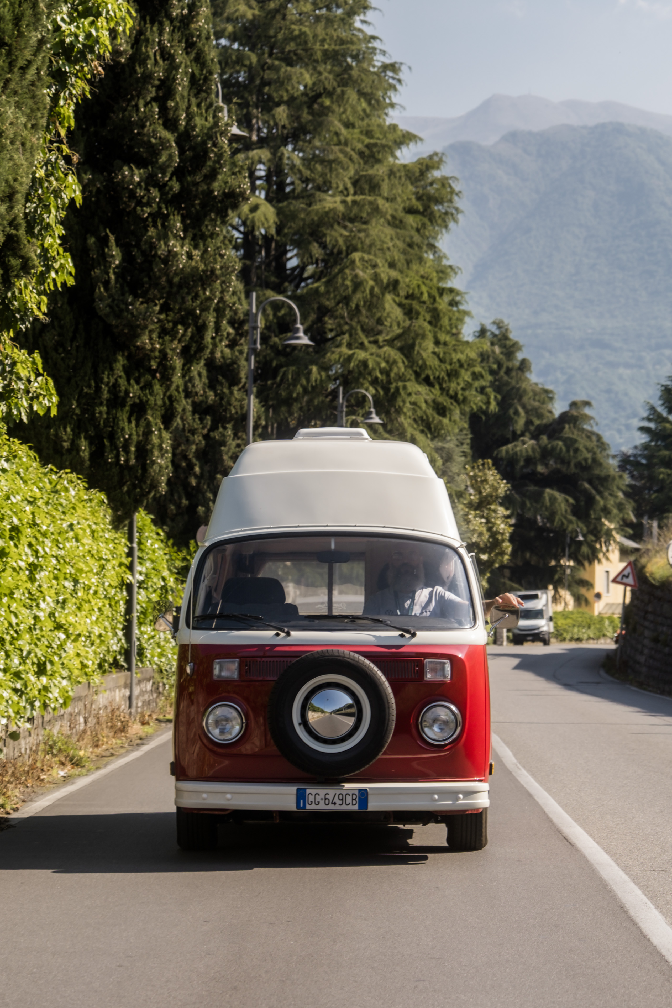Un classico camper VW rosso e bianco in viaggio su strada, tra alberi rigogliosi e montagne mozzafiato.