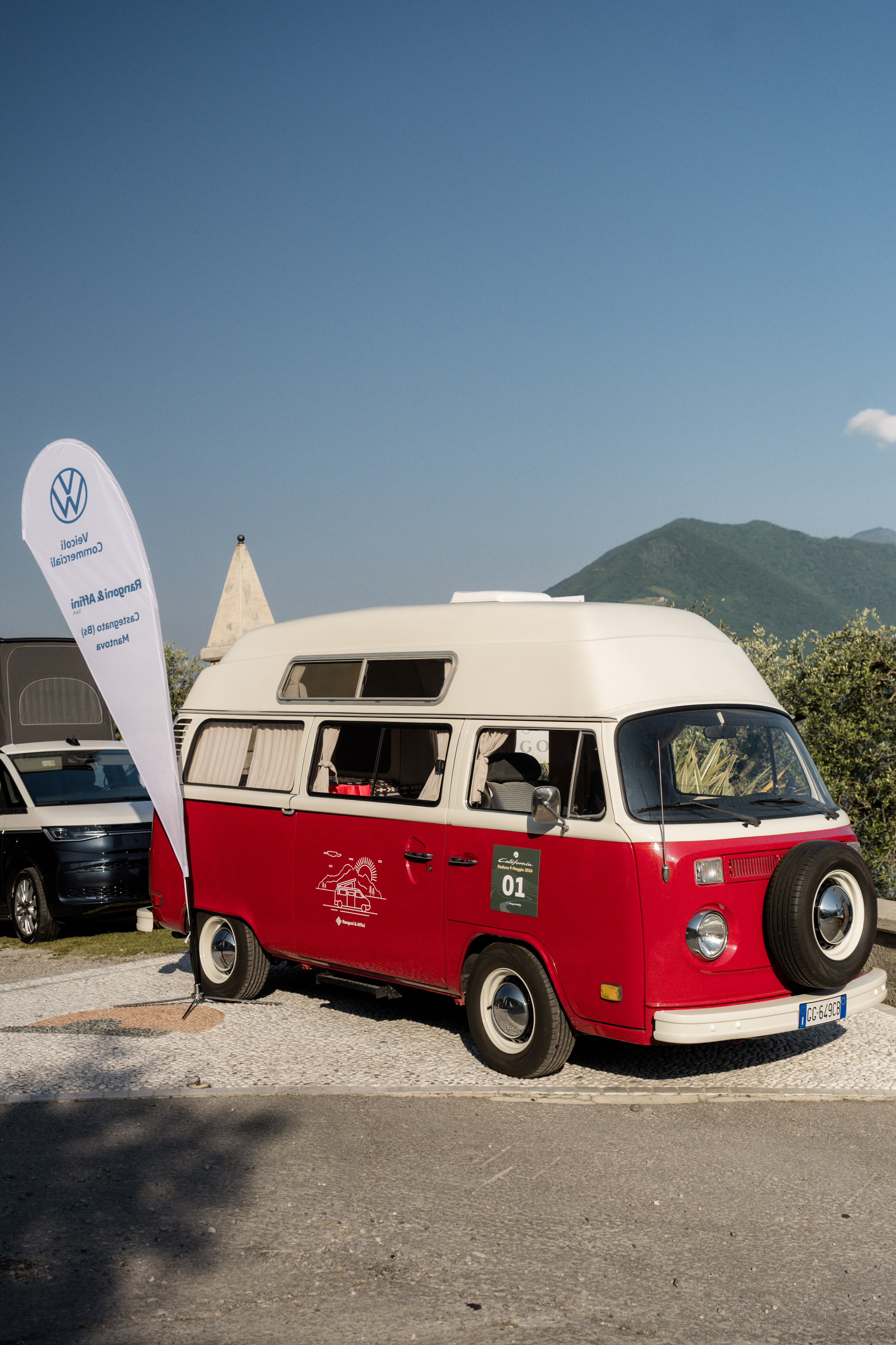 L'iconico camper Volkswagen rosso e bianco, numero 01, a un raduno, con le montagne sotto un cielo azzurro.