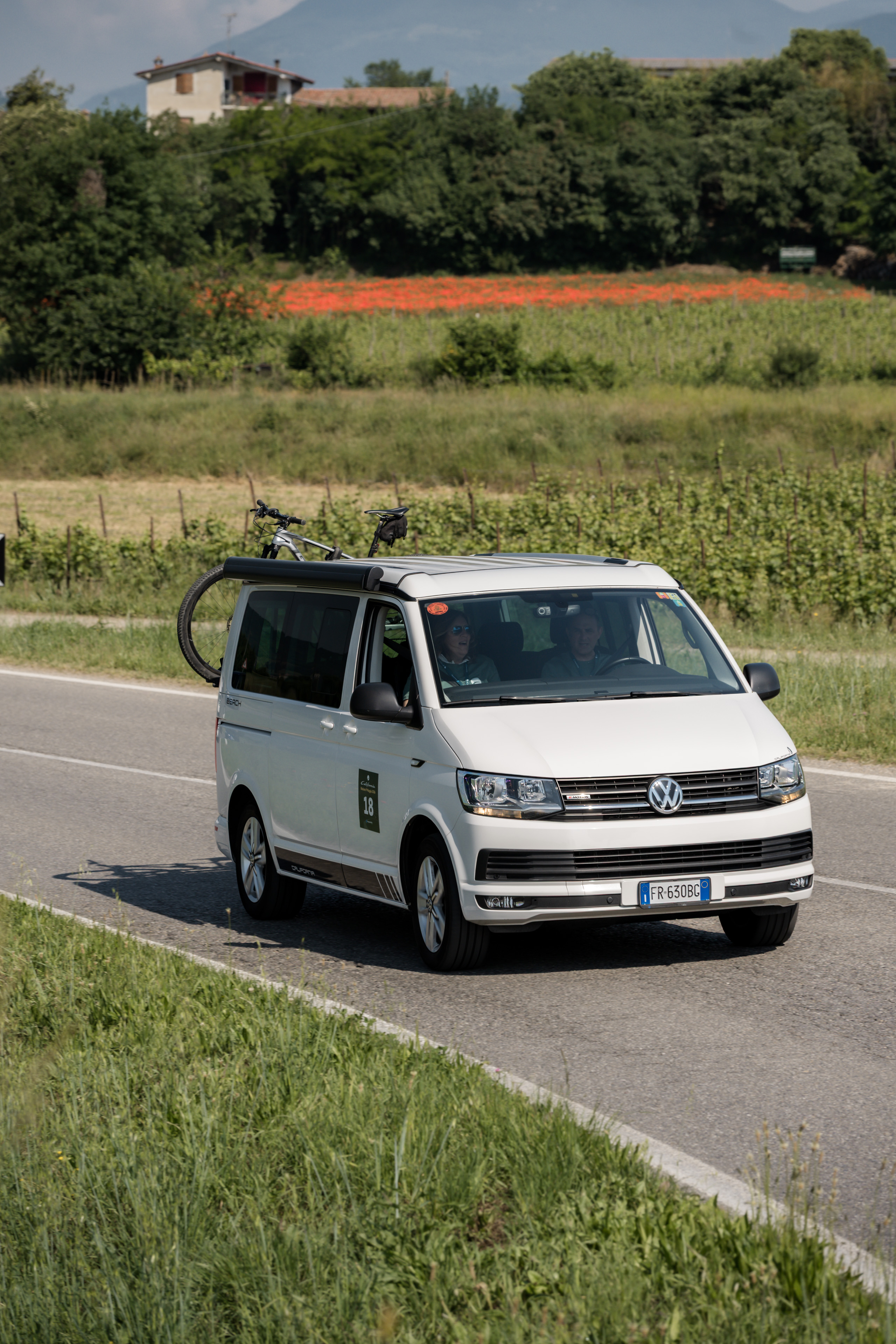 Volkswagen California van with bici su strada panoramica in Italia, tra campi di papaveri e vigneti.