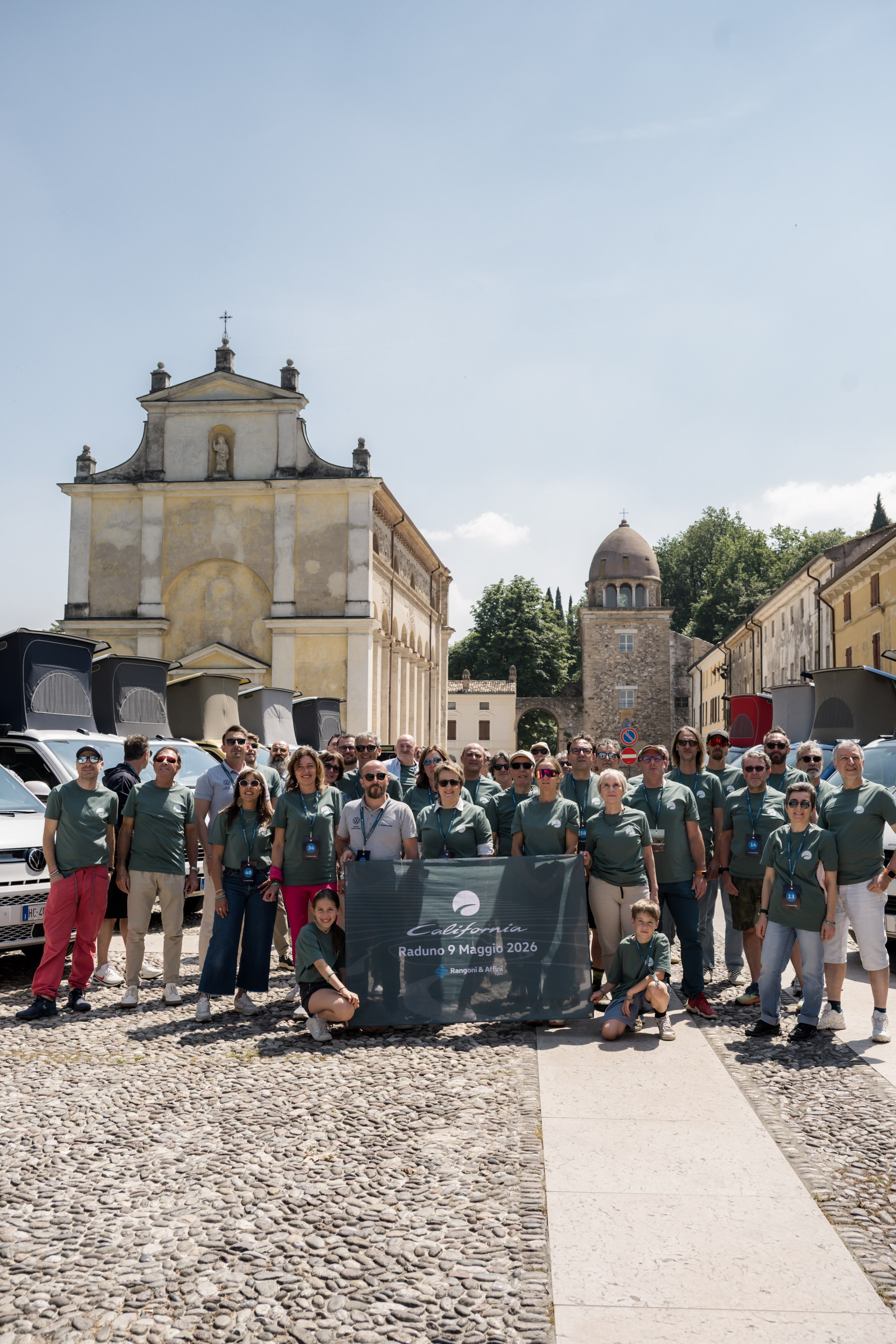 Gruppo di persone, inclusi bambini, al 'California Raduno 9 Maggio 2026', con camper e architettura storica sullo sfondo.