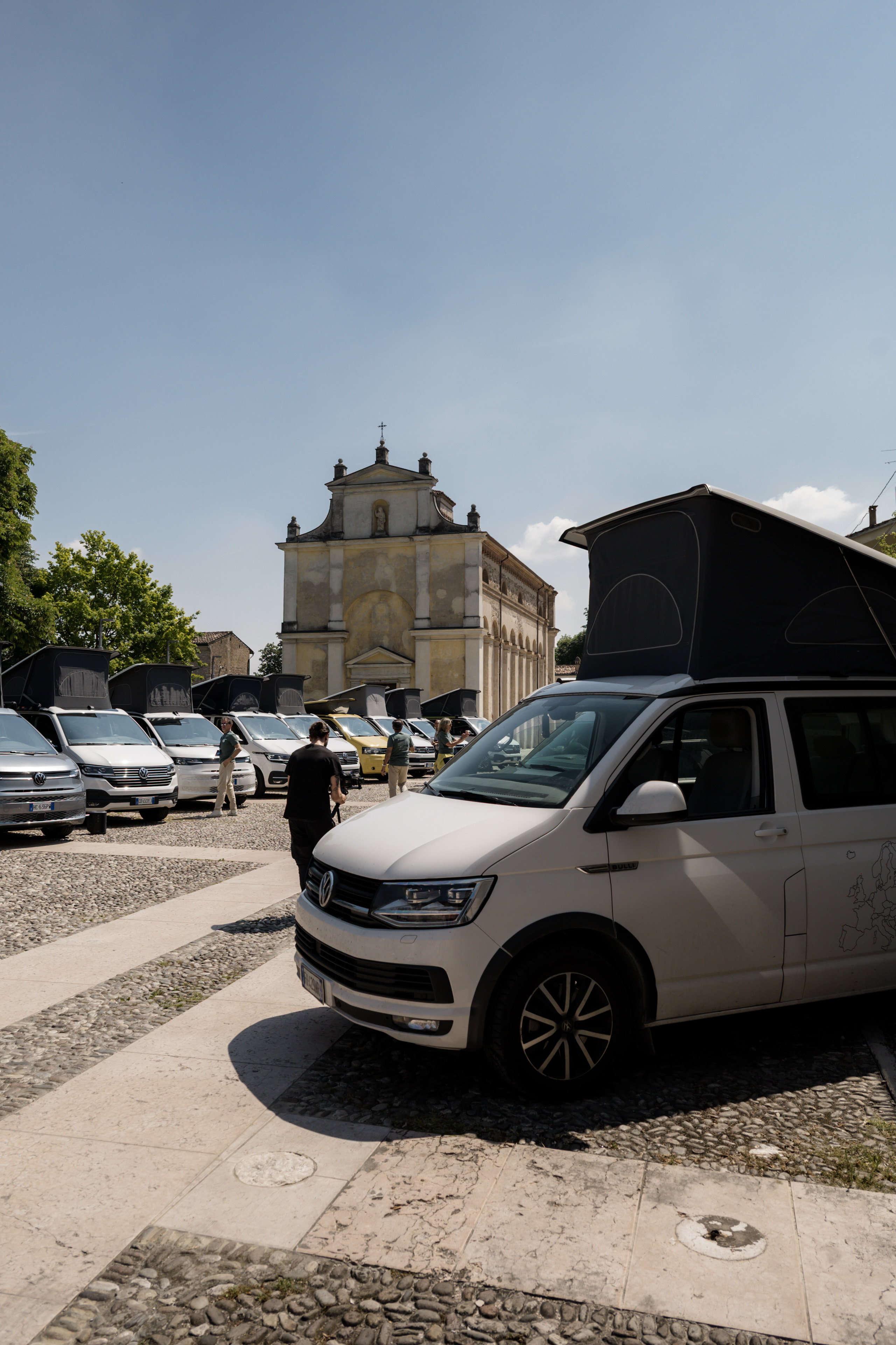 Una carrellata di camper van con tetto a soffietto parcheggiati in una piazza acciottolata, con una chiesa storica sullo sfondo.