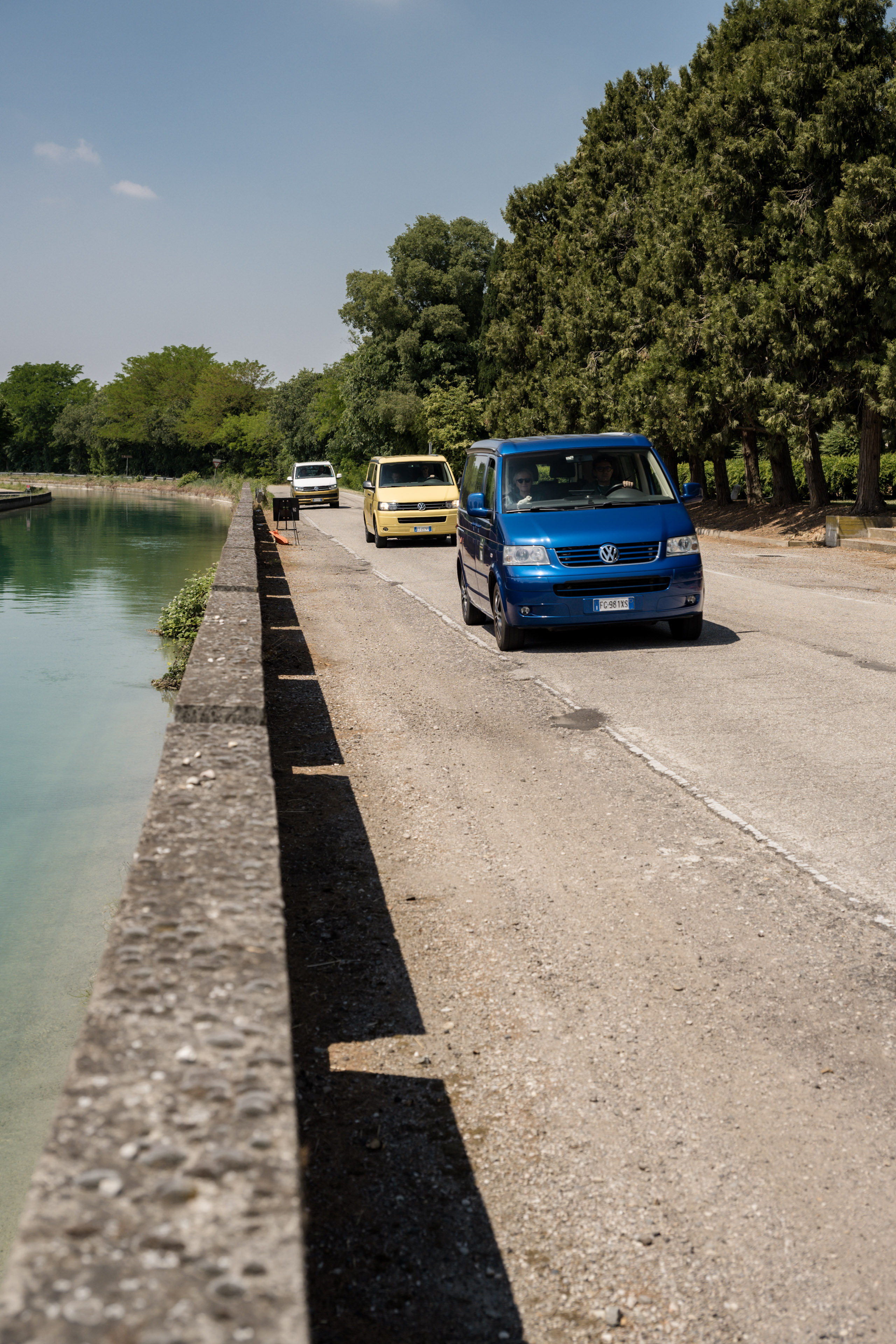 Convoglio di furgoni VW colorati su strada assolata, con canale e alberi sullo sfondo.