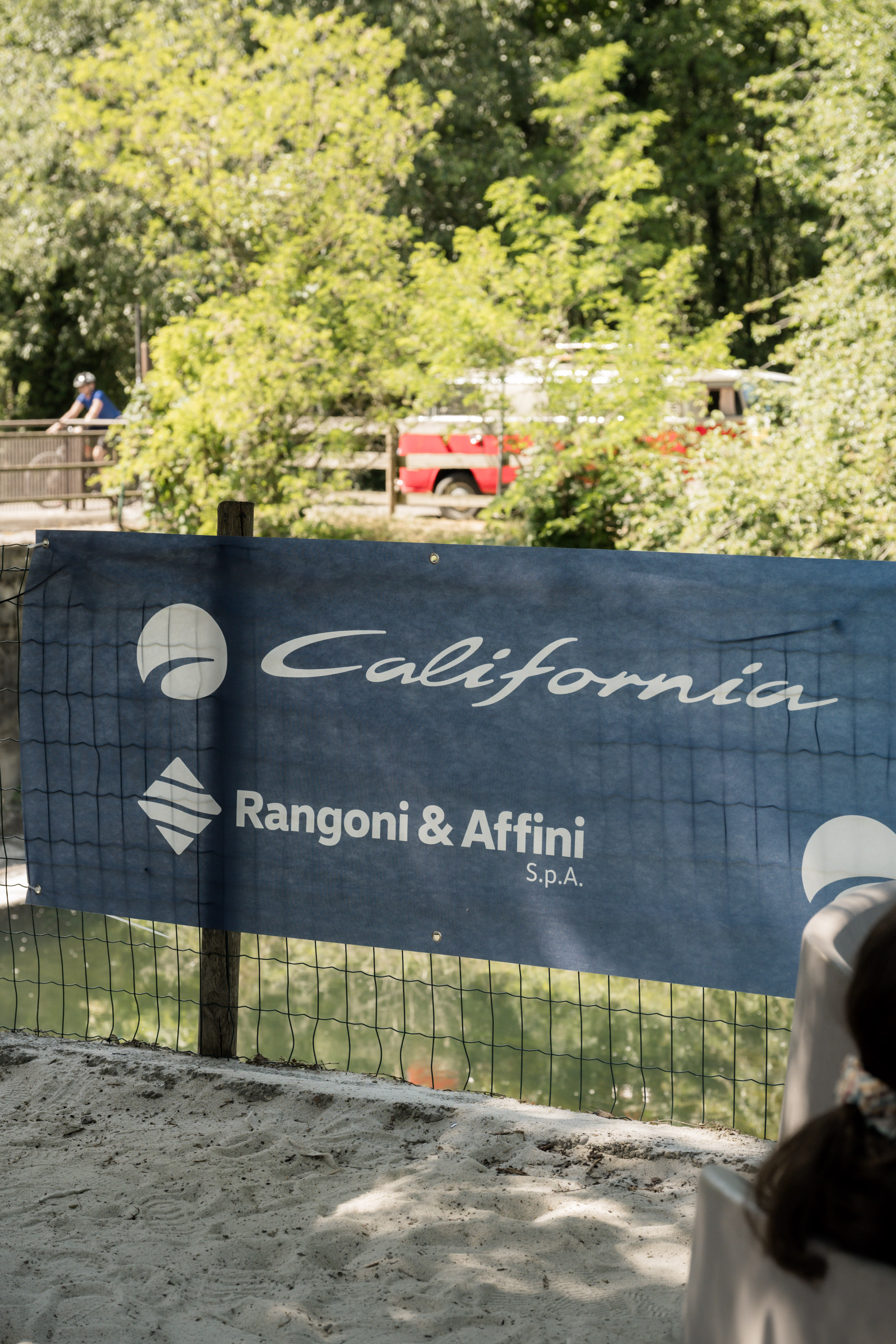 Banner "California Rangoni & Affini" su recinzione, con sabbia, acqua, vegetazione e un veicolo rosso in lontananza.