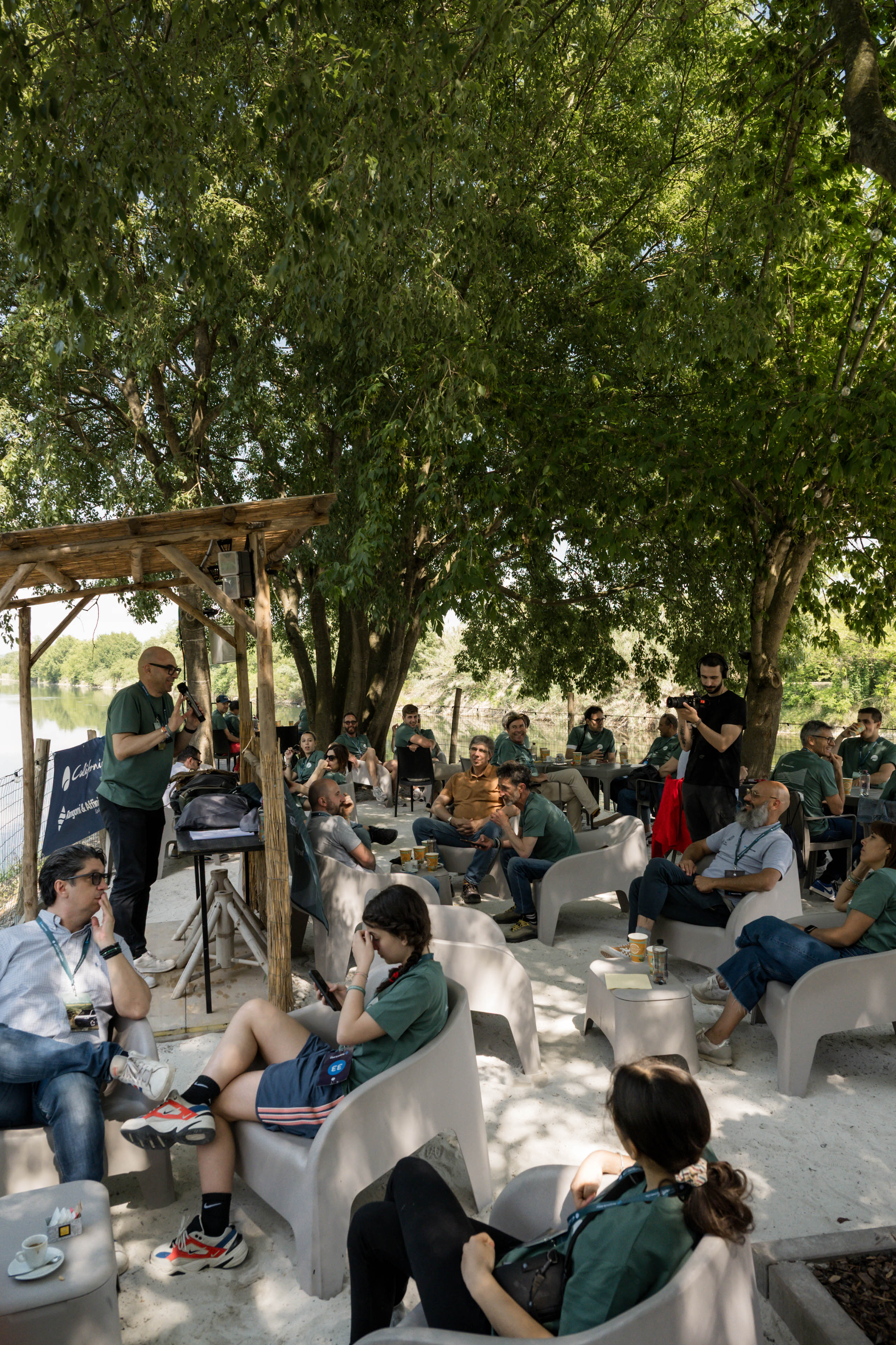 Conferenza informale all'aperto in riva al fiume, con oratore e pubblico rilassato sotto gli alberi.