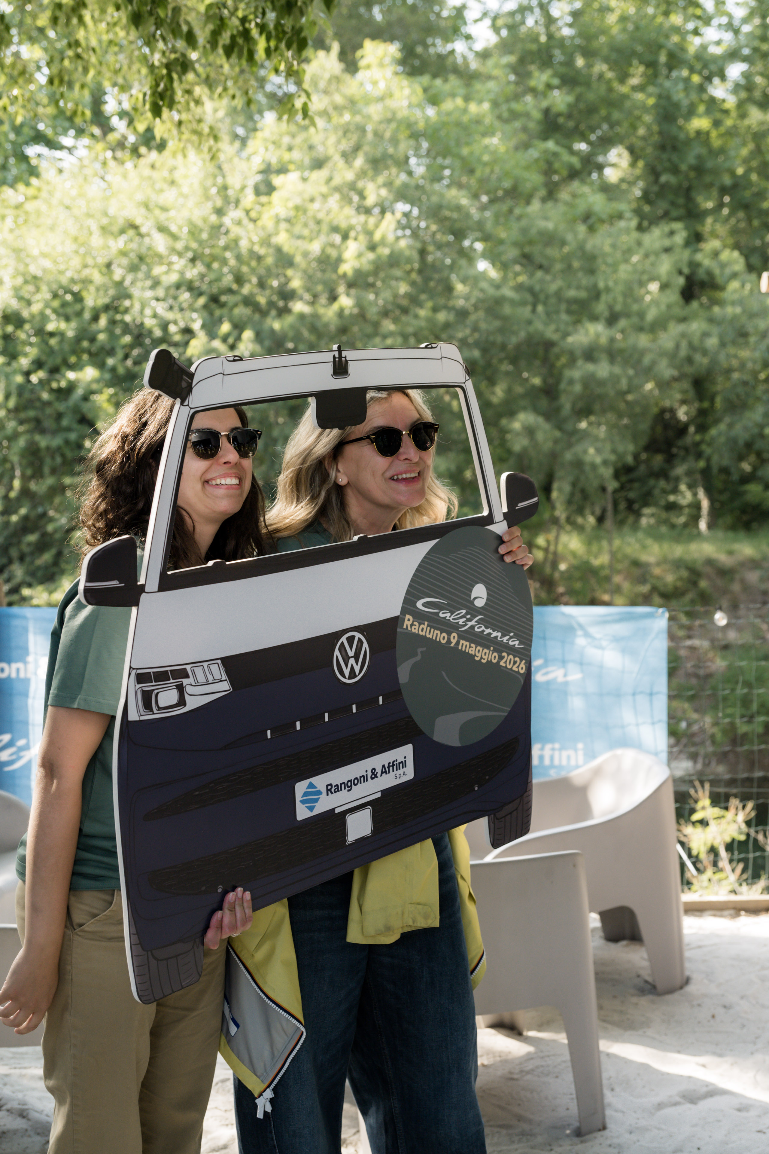 Due donne sorridenti si divertono posando con un cutout di Volkswagen California, pronti per il raduno del 2026!