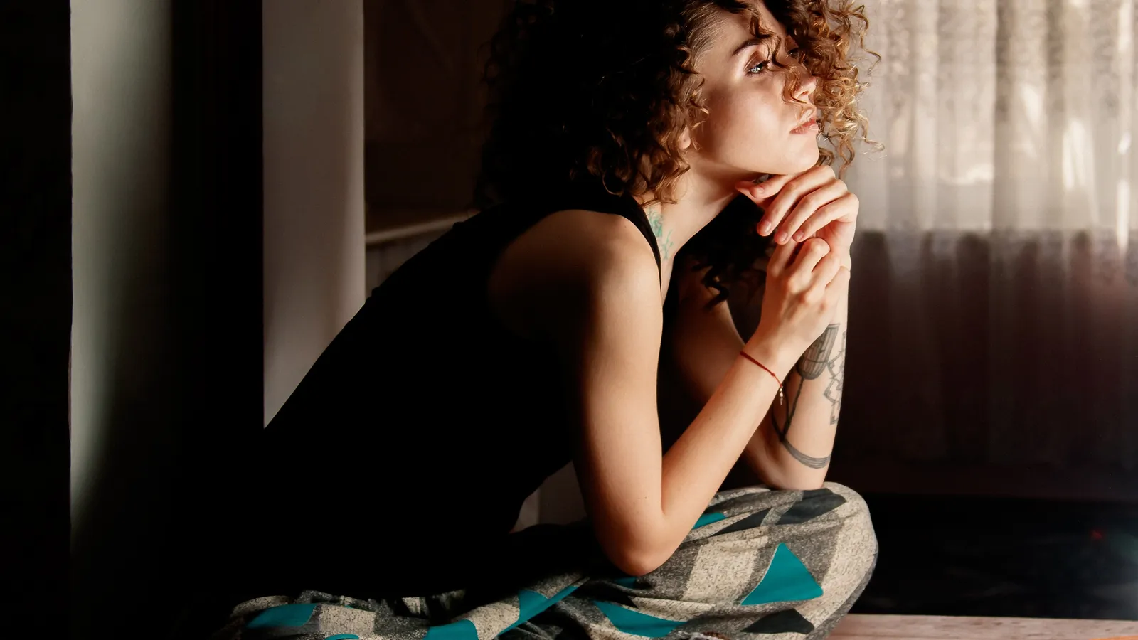 Woman sitting cross-legged on a bed by a window, resting her chin on her hands