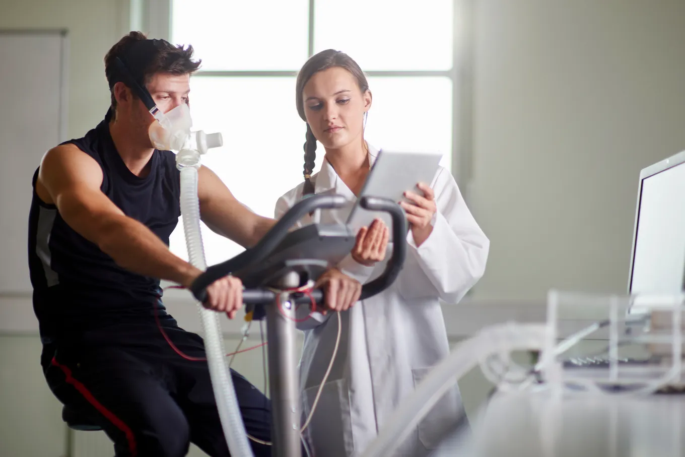 Man pedaling a stationary bike wearing a metabolic cart mask while a technician in a lab coat records the readings