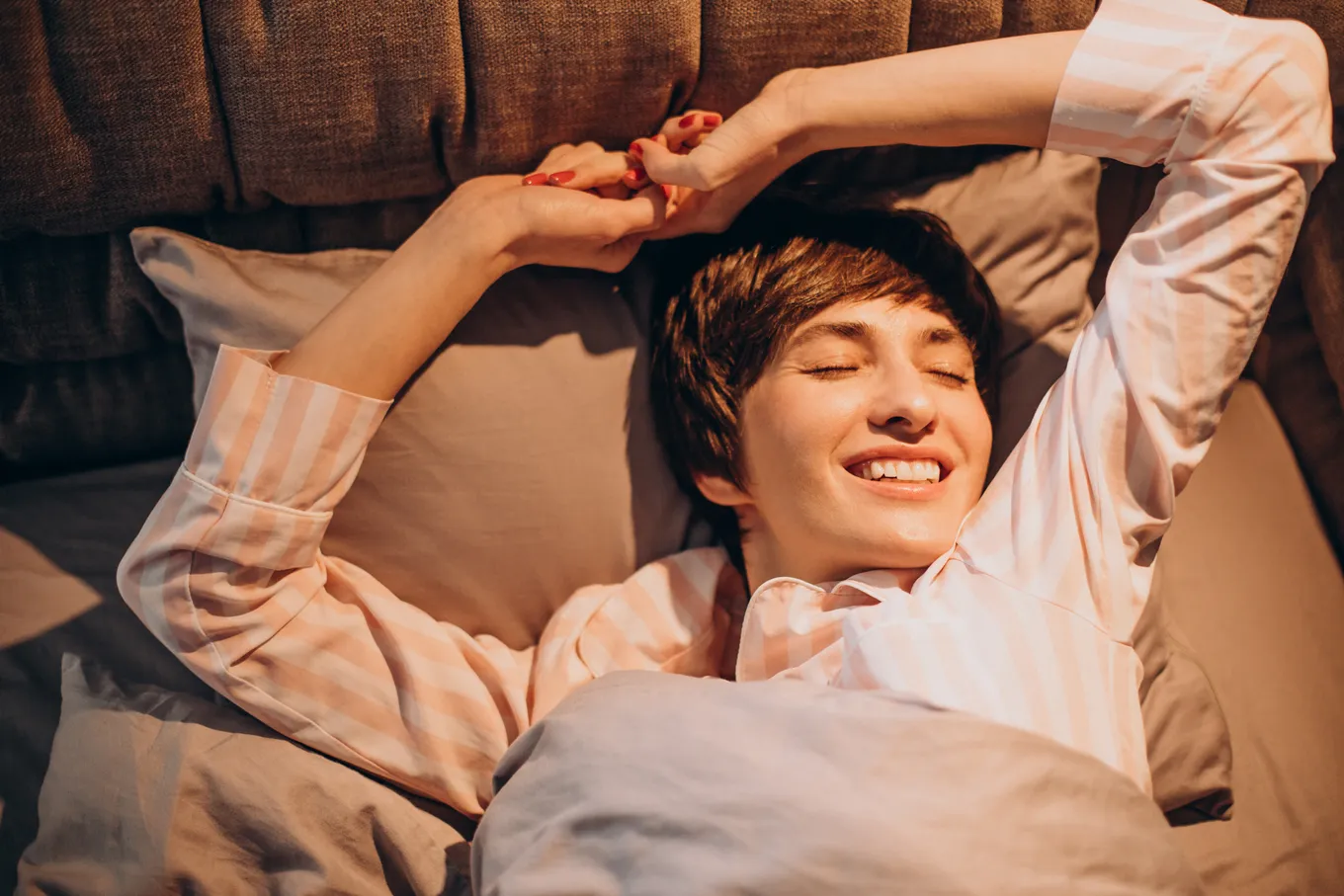 Woman in striped pajamas waking in bed, arms stretched above her head in morning light