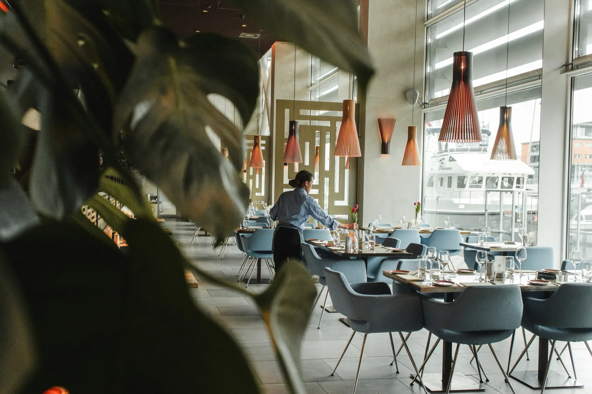 A bright restaurant dining room with set tables and copper pendant lights beside tall windows