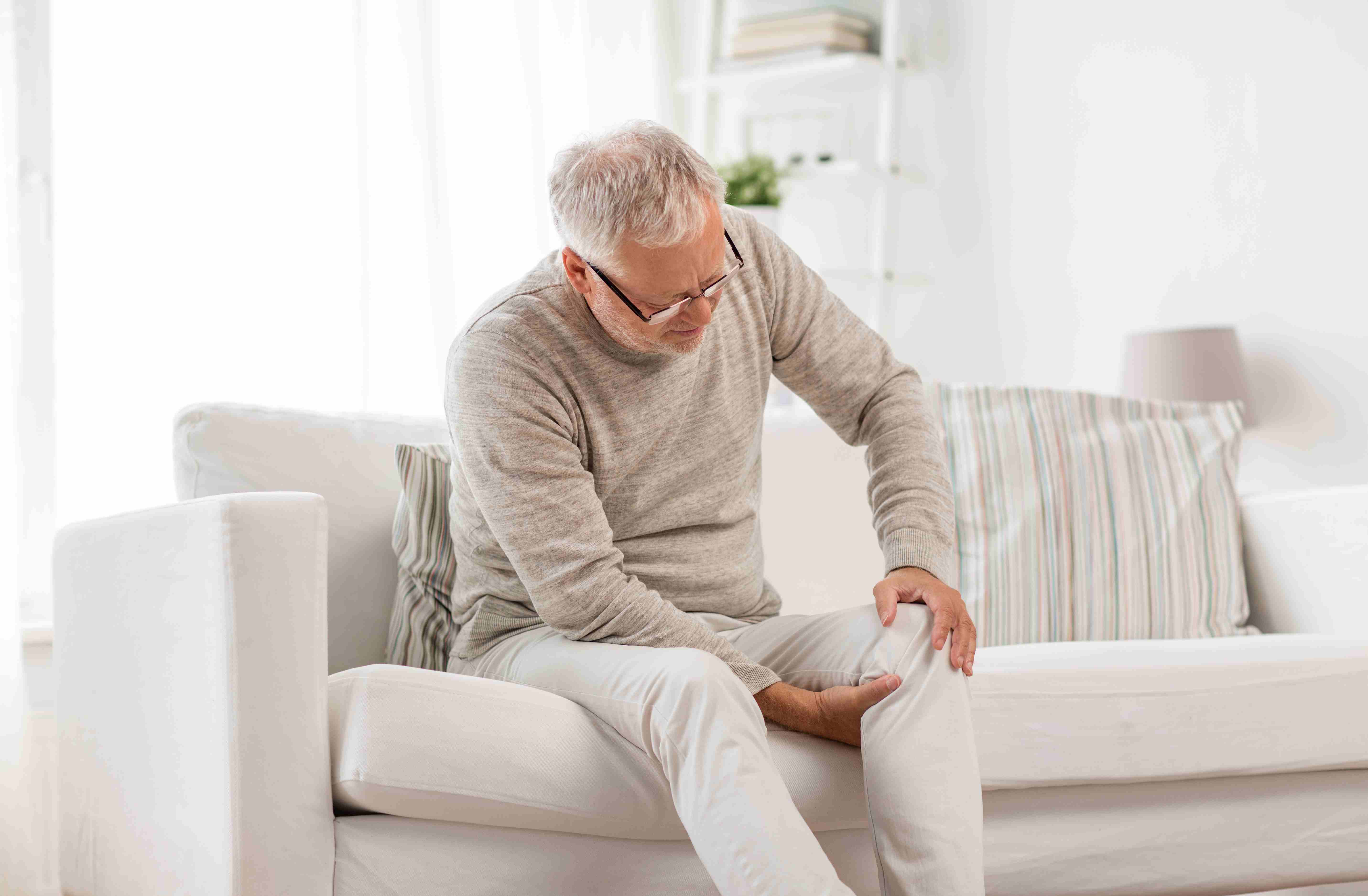 Senior man sitting on a couch holding his knee in pain, showing signs of chronic joint discomfort before knee replacement surgery.