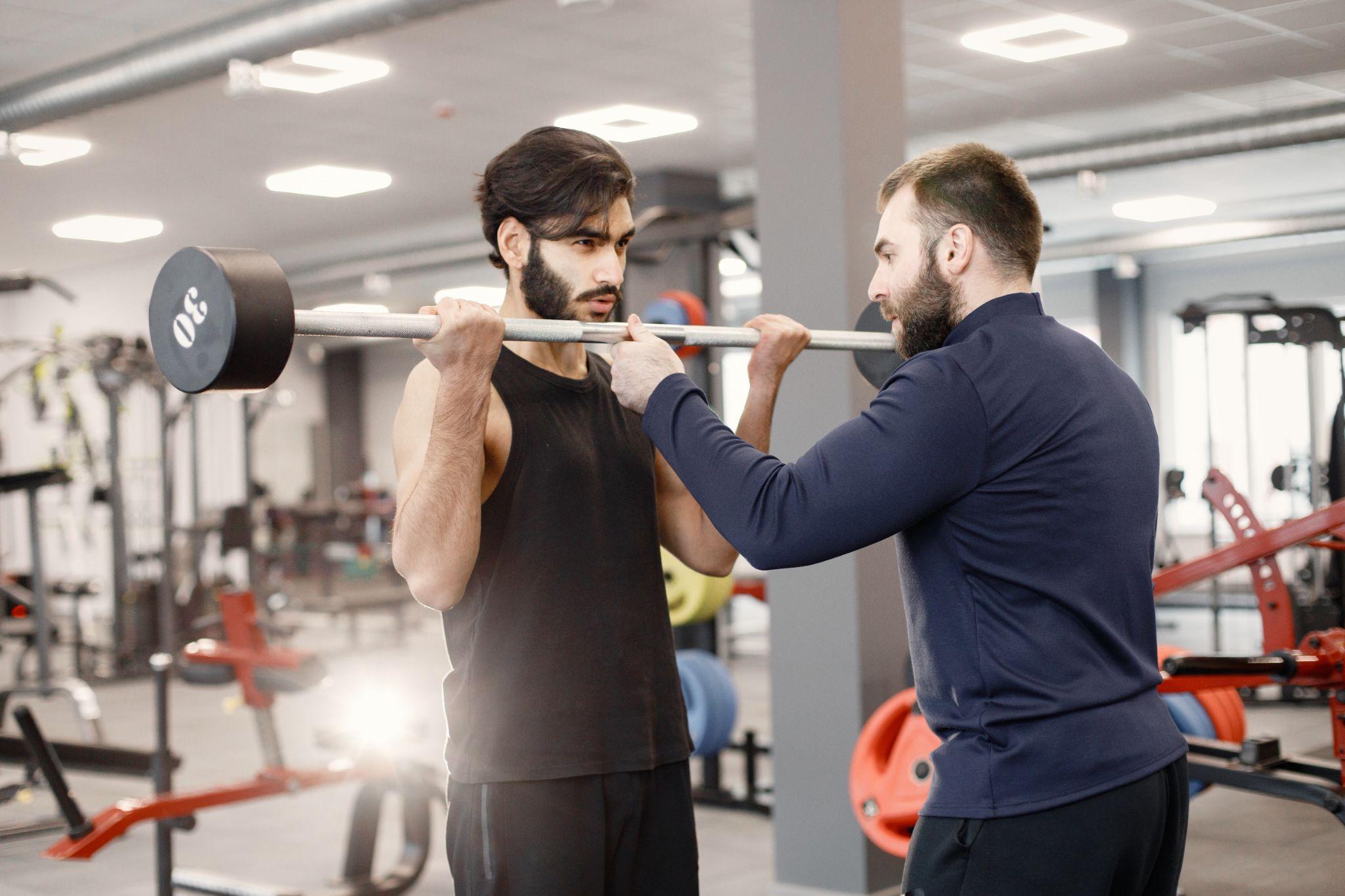 A man is performing a barbell exercise under the close supervision of a gym trainer. Both individuals appear focused, with the trainer guiding the proper lifting technique.