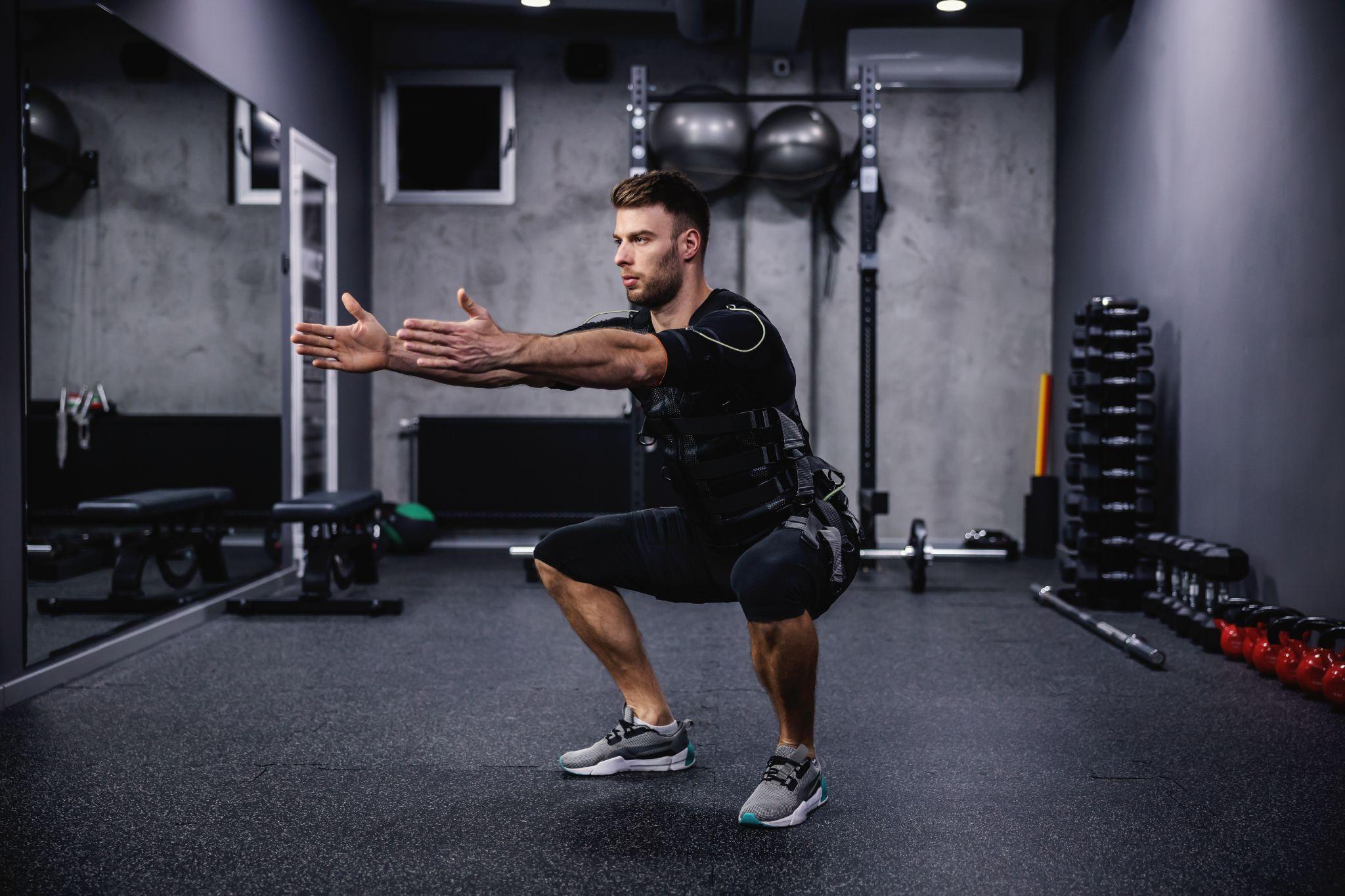 A man in a gym performs a squat. The dimly lit fitness room includes weights and equipment in the background, highlighting strength and conditioning exercise.