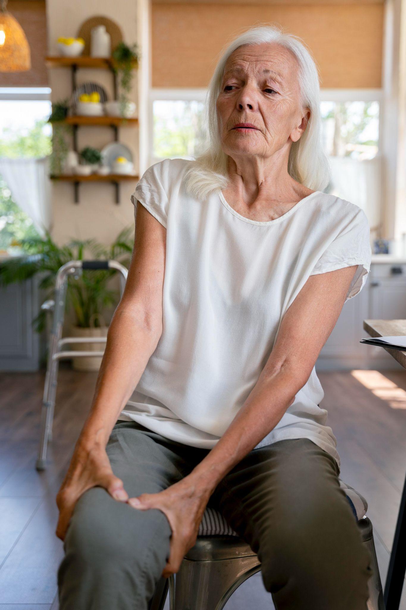 An elderly woman with white hair sits on a chair inside her home, holding her knee with visible discomfort. Her facial expression suggests knee pain or arthritis symptoms. The background shows a bright kitchen with plants and shelves, giving a natural, homely environment.