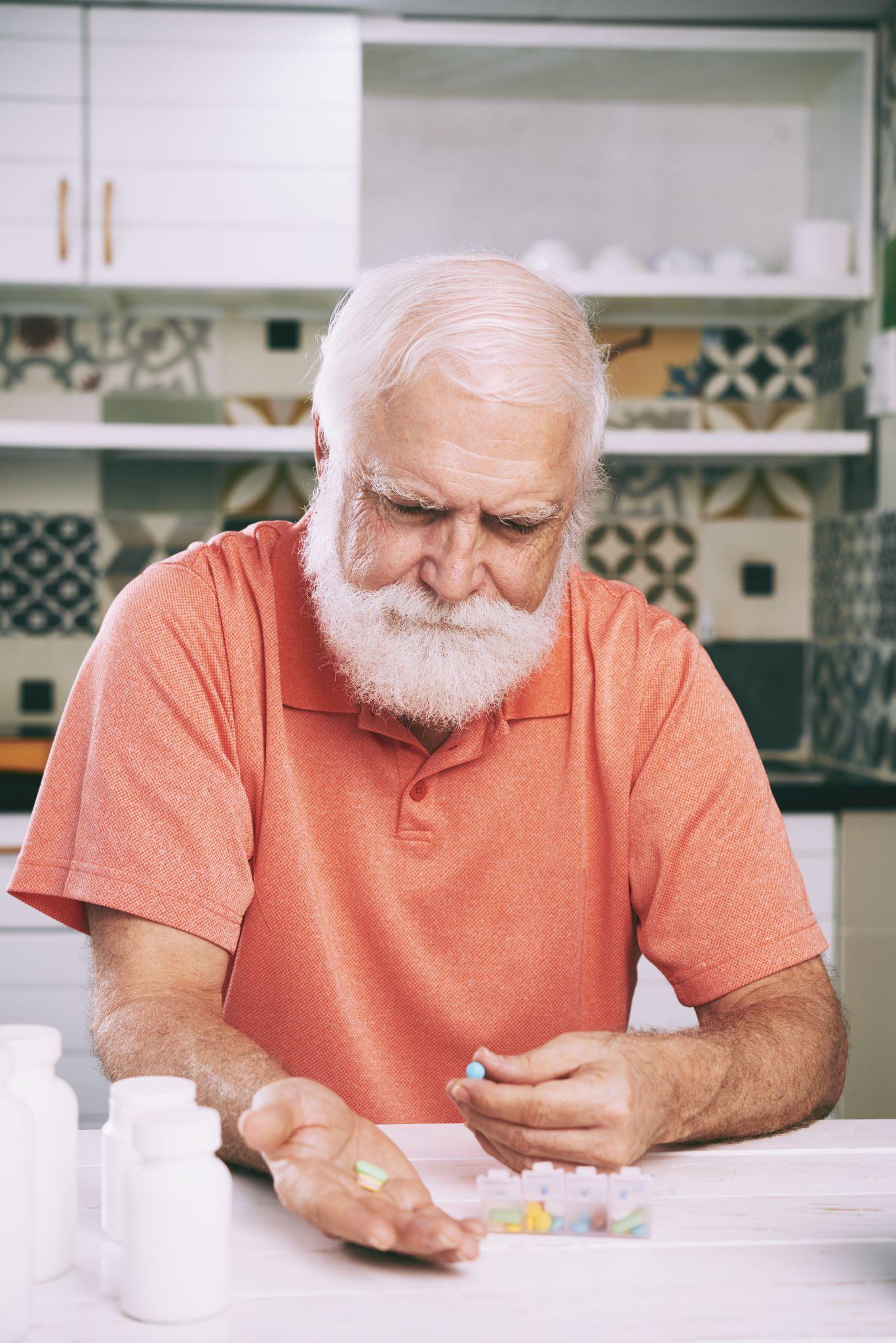 An older man with white hair and a beard sits at a table in his kitchen, organizing pills in a medication box. He appears focused while holding tablets in one hand. Multiple medicine bottles are placed on the table, indicating ongoing treatment or management of a chronic health condition like arthritis.