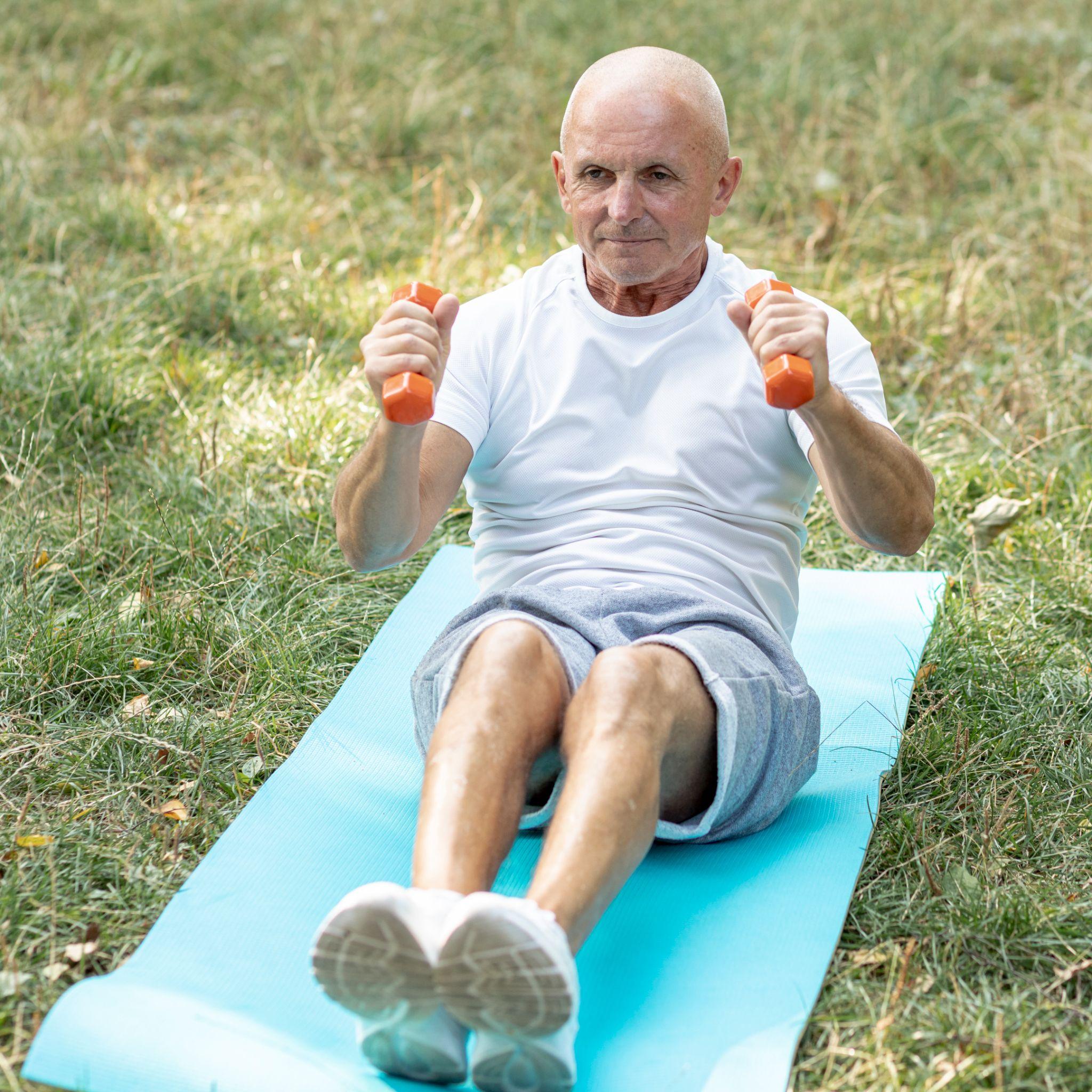 An elderly man exercises outdoors while sitting on a yoga mat and lifting small dumbbells. He appears focused and active, showcasing strength training as part of a healthy lifestyle.